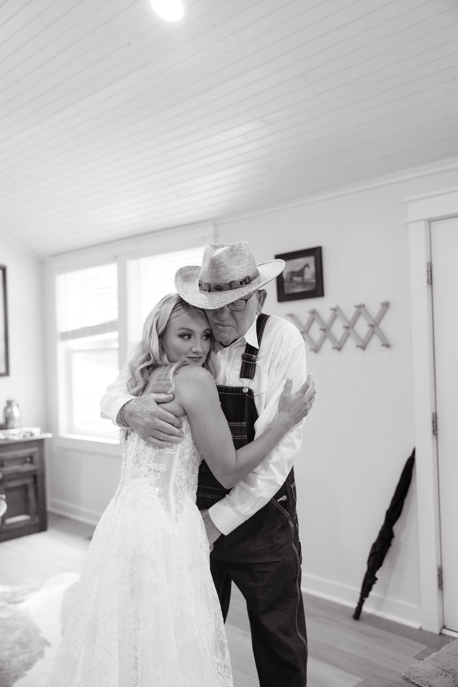 Black and white photo of bride in lace gown embracing man in cowboy hat and overalls indoors.