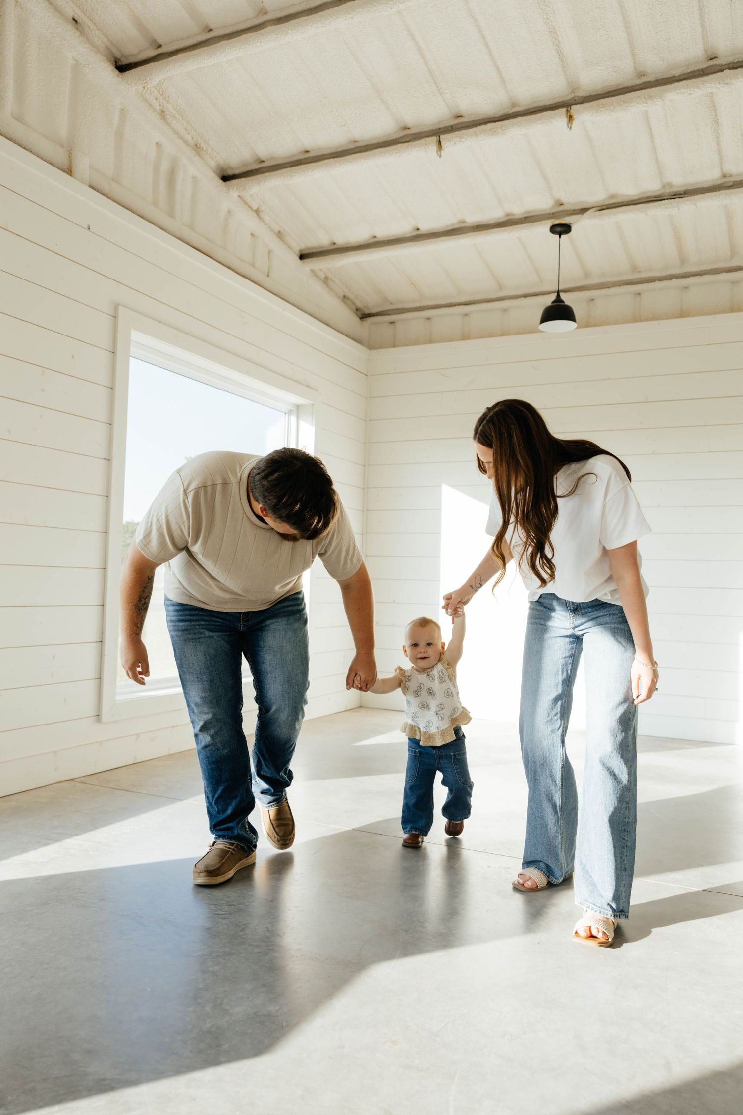 Young parents helping their baby take first steps inside bright white modern home interior.