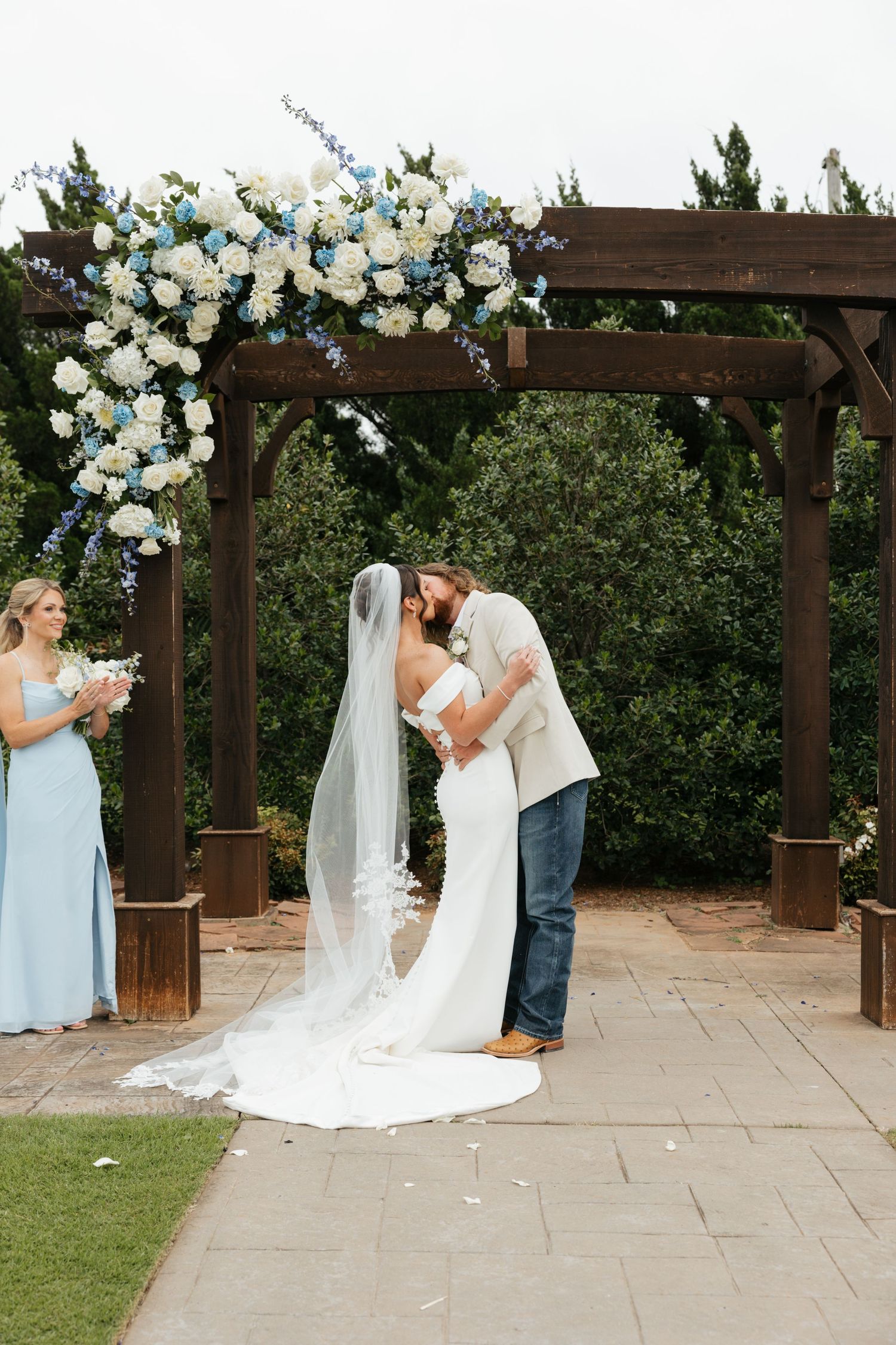 Bride and groom share first kiss under wooden pergola adorned with blue and white floral arrangement.