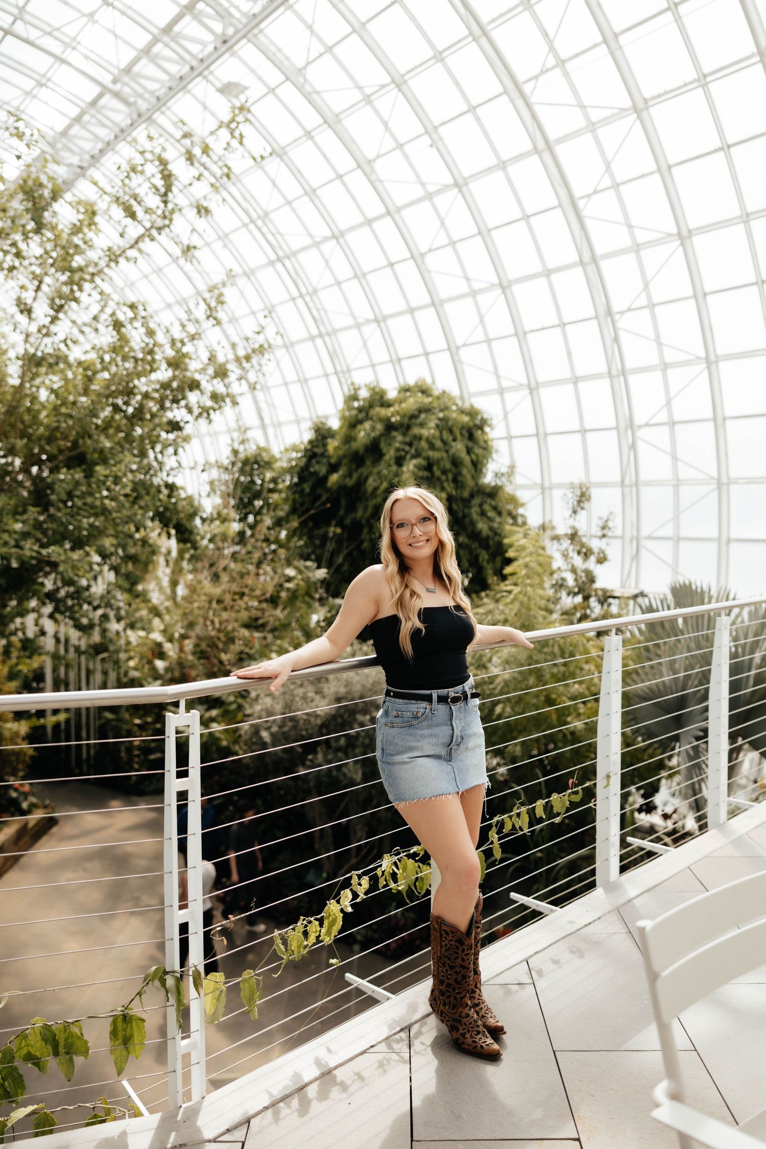 Blonde woman in black top, denim skirt, and leopard boots posing in a greenhouse conservatory.