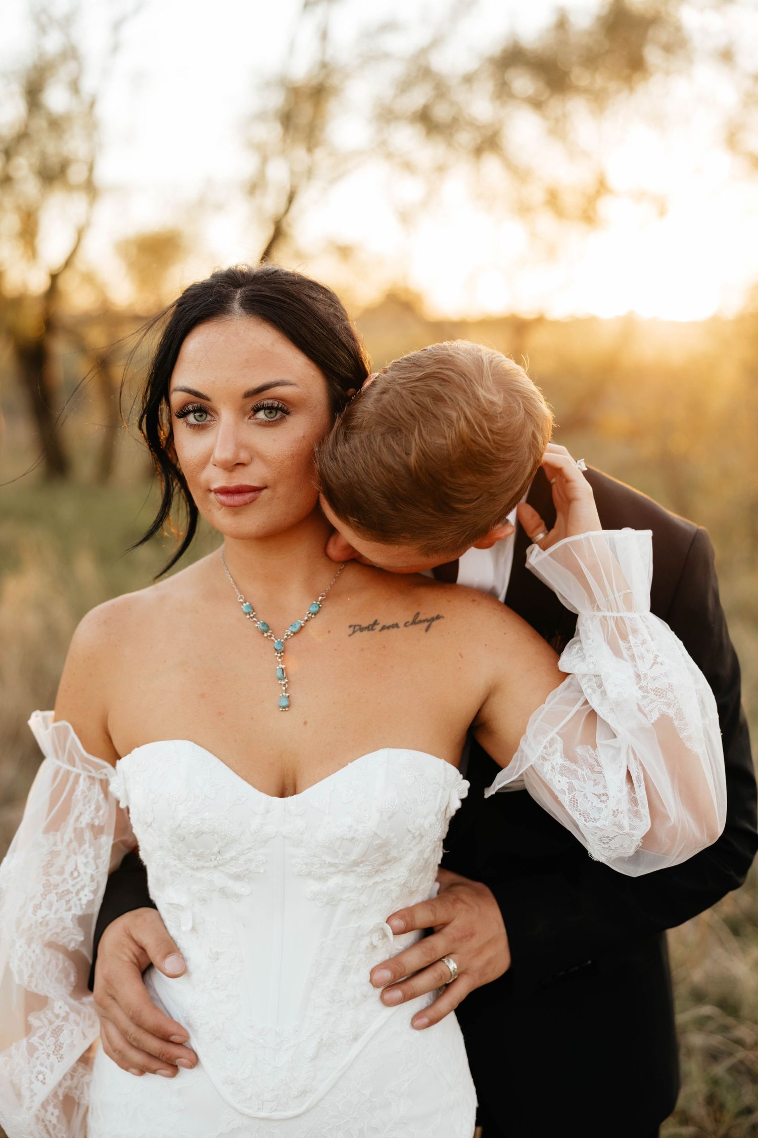 Bride in white lace gown with turquoise necklace embraced by groom at golden hour outdoor wedding.