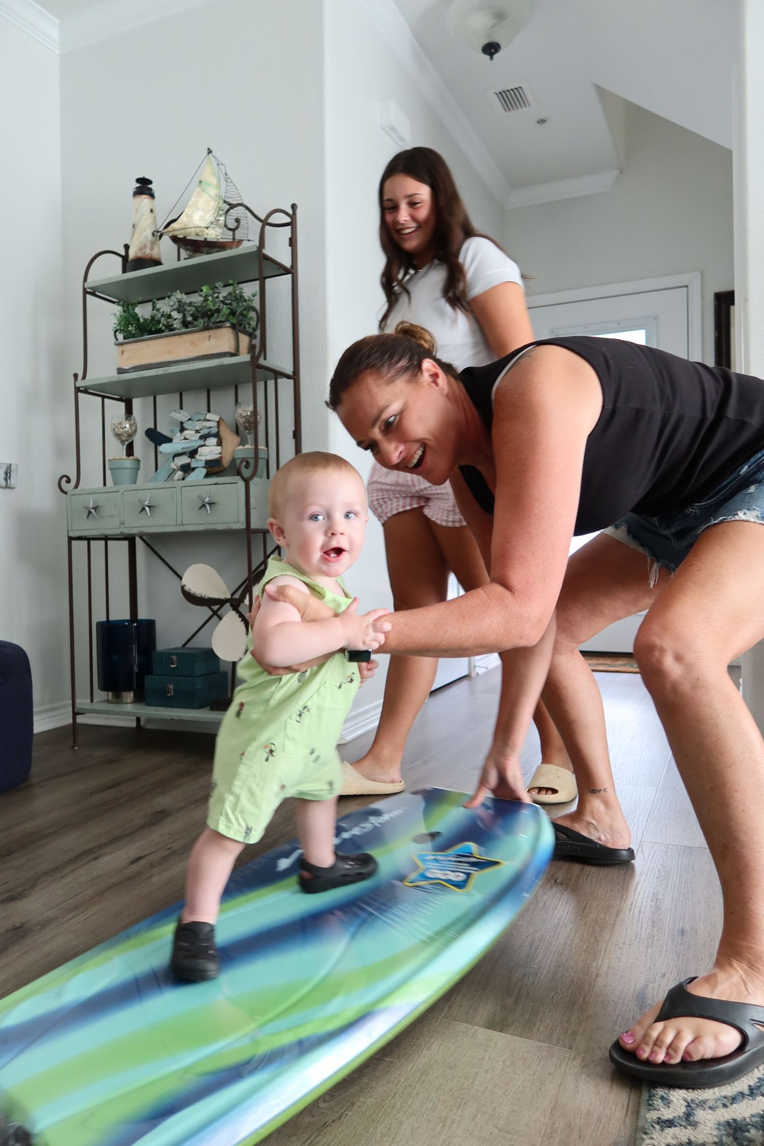 Woman helps excited baby stand on surfboard indoors while teenager watches and smiles.
