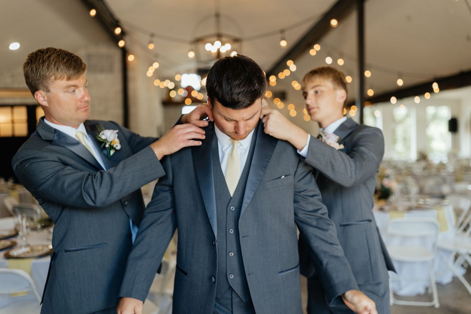 Groom being helped by two groomsmen adjusting his suit collar inside decorated wedding venue.