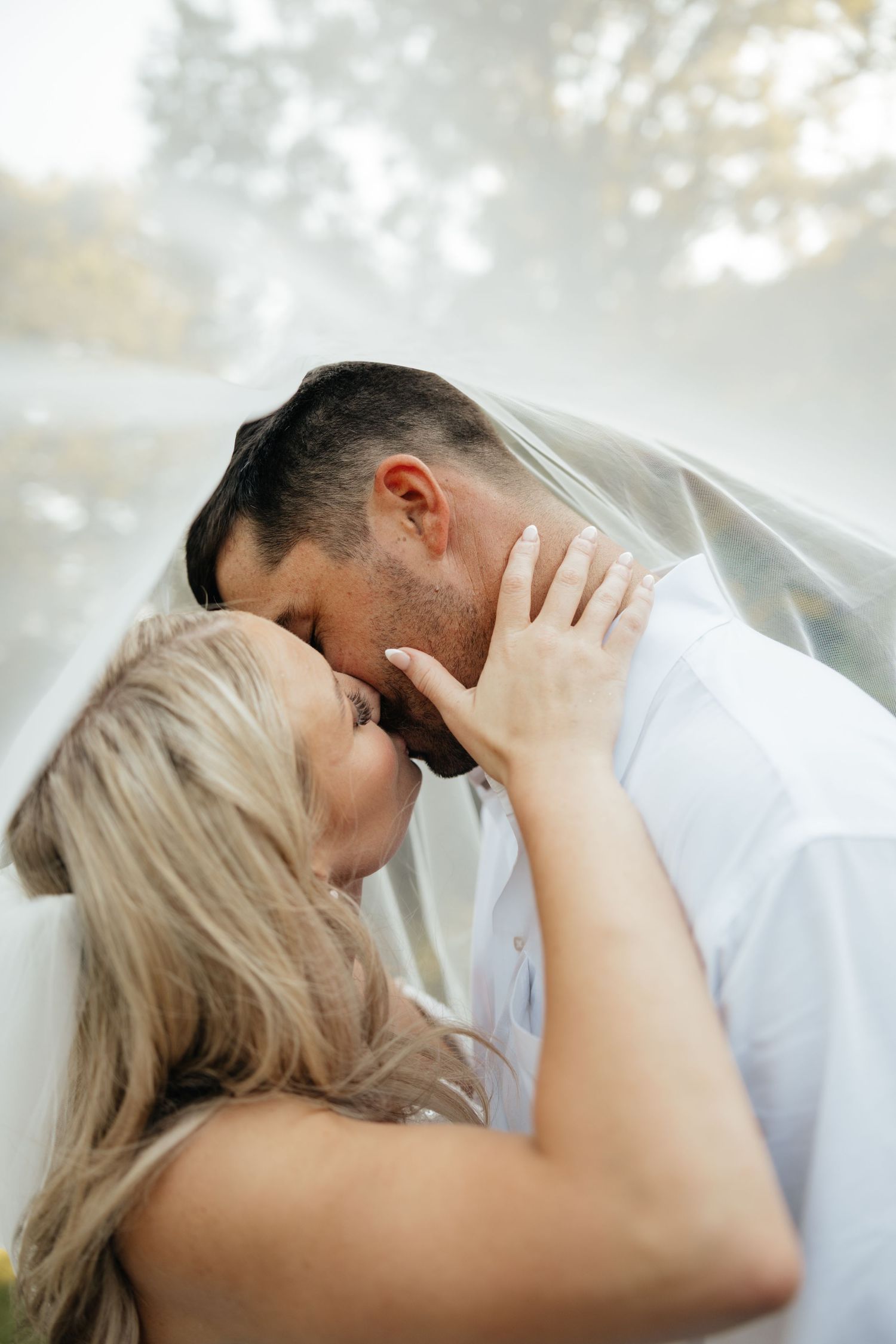 Bride and groom kissing intimately under sheer veil, brides hand gently cradling grooms face outdoors.