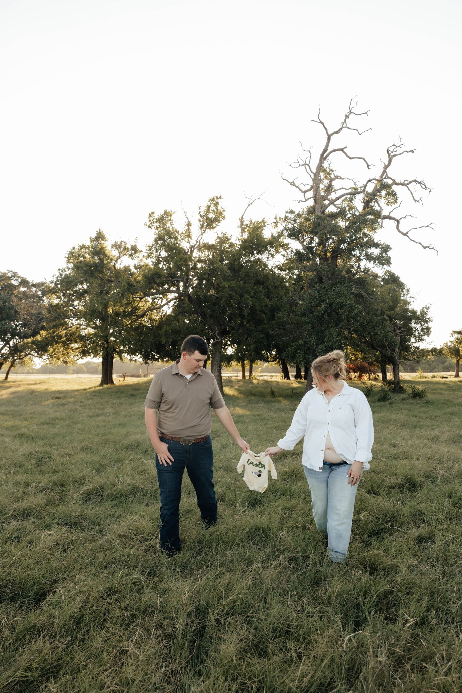 Expectant couple walking through green field at sunset holding tiny baby onesie between them.