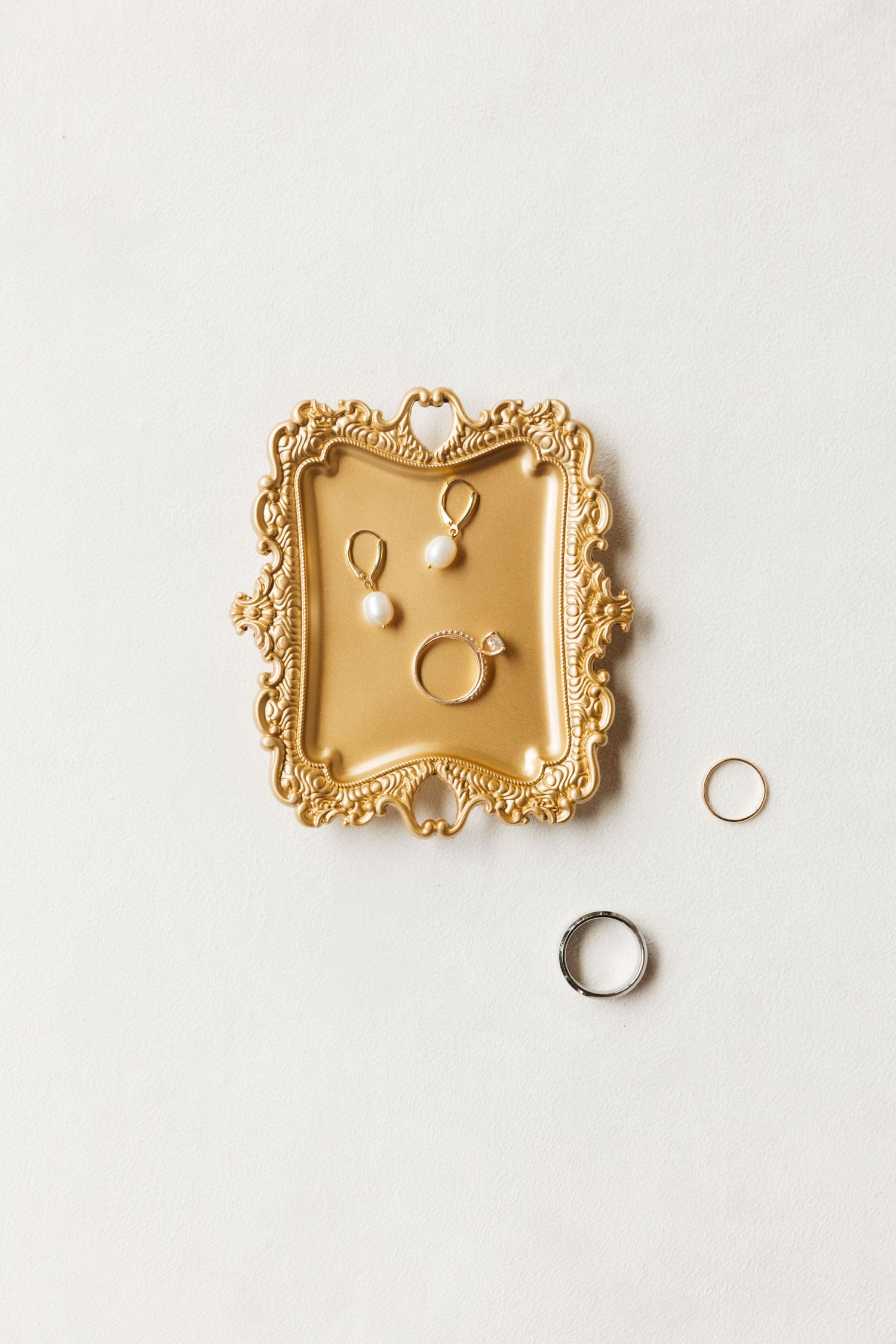 Gold ornate tray displaying pearl earrings, diamond engagement ring, and wedding bands on white surface.