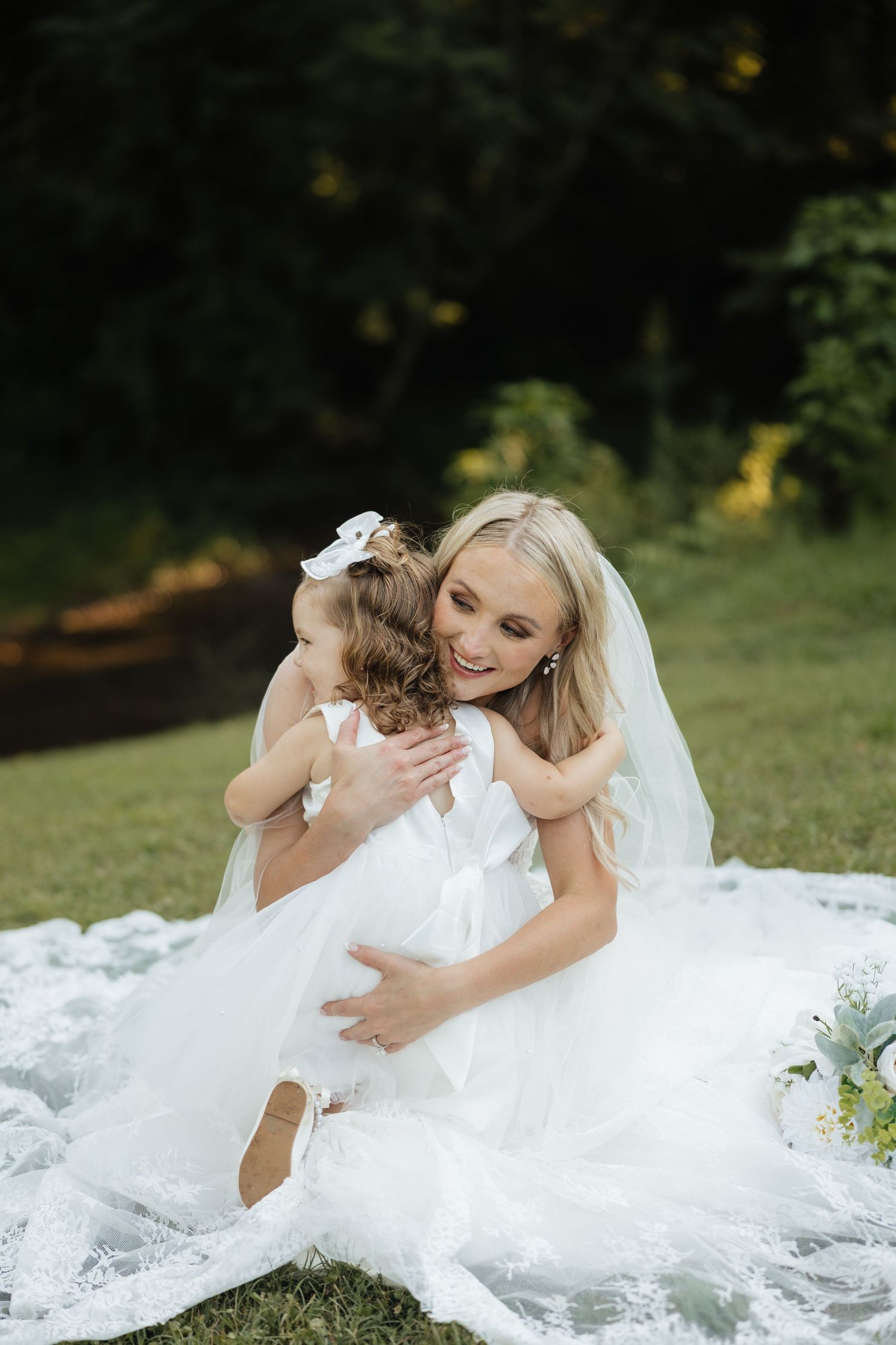 Bride in white lace gown and veil sits on grass hugging young flower girl in white dress outdoors.