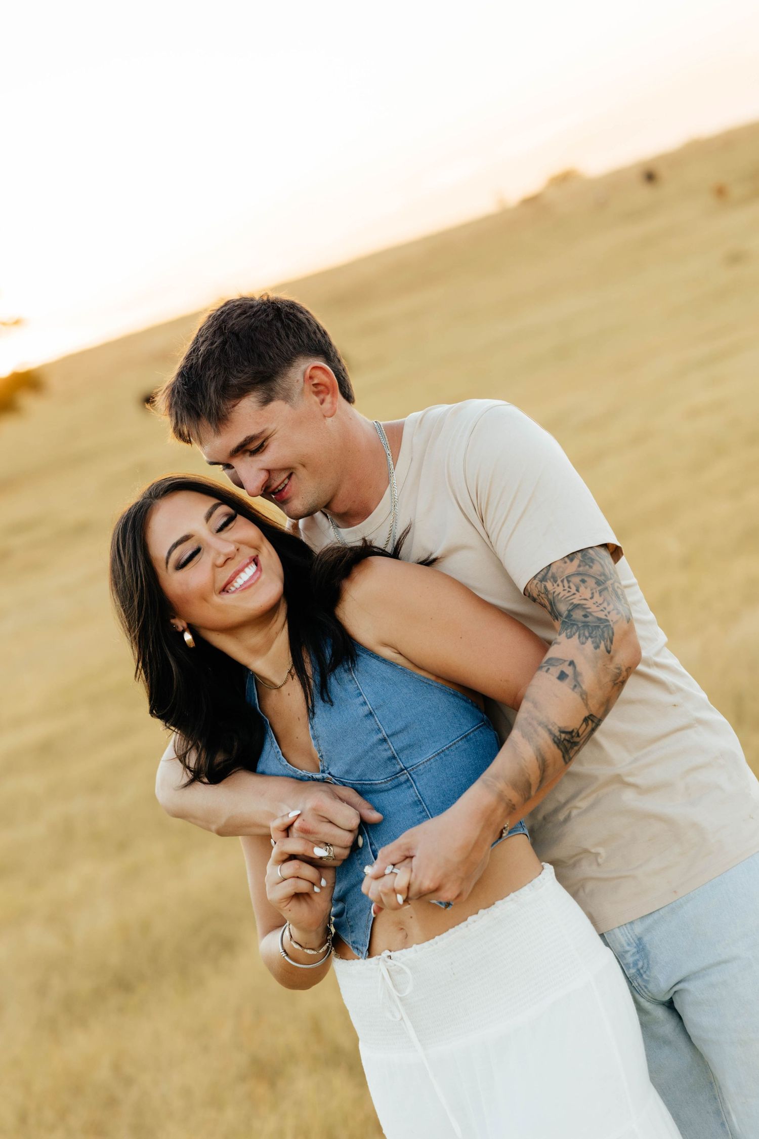 Couple embracing at sunset in golden field, woman laughing as man hugs her from behind.
