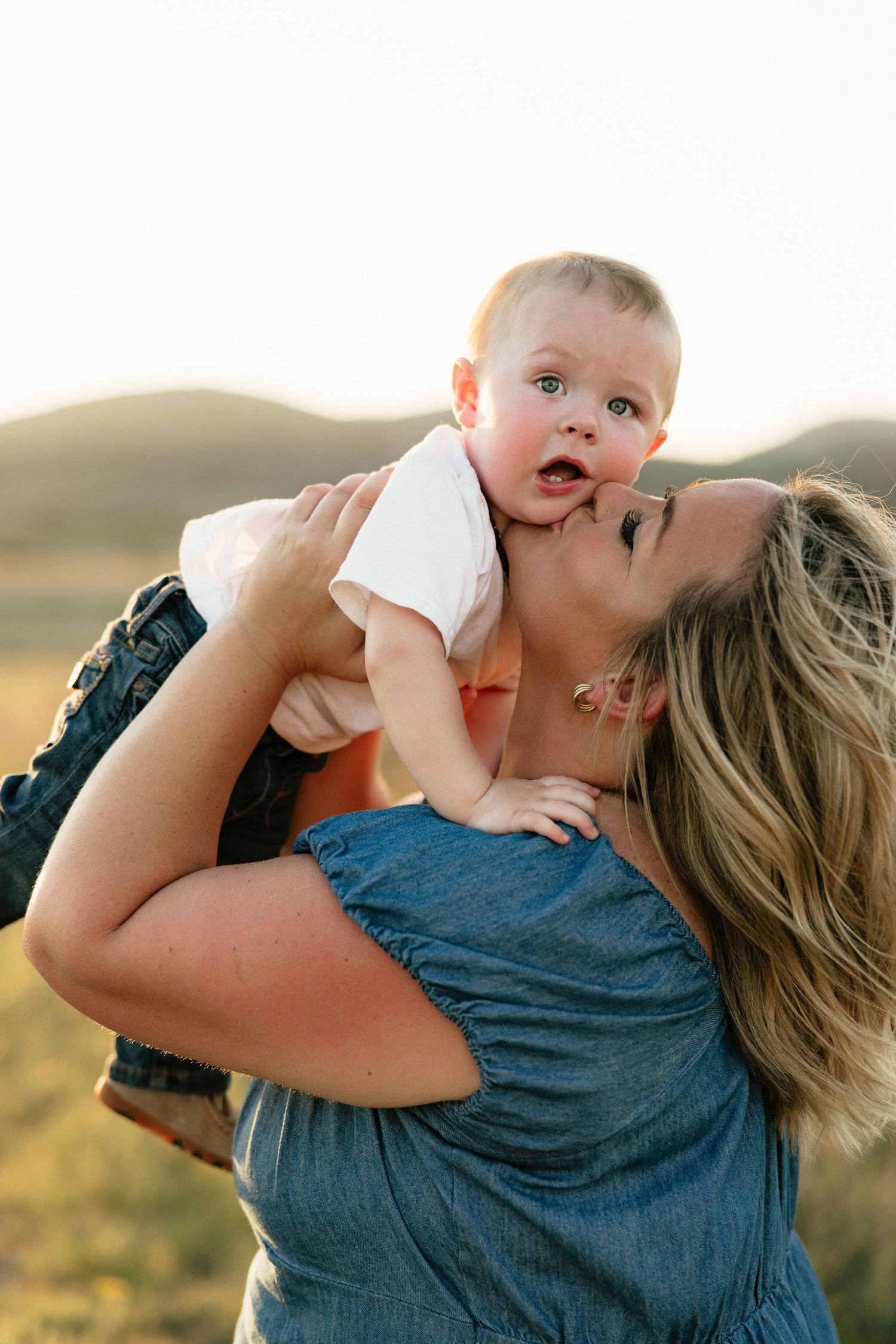 Mother kissing and lifting smiling baby outdoors at golden hour with mountain backdrop.