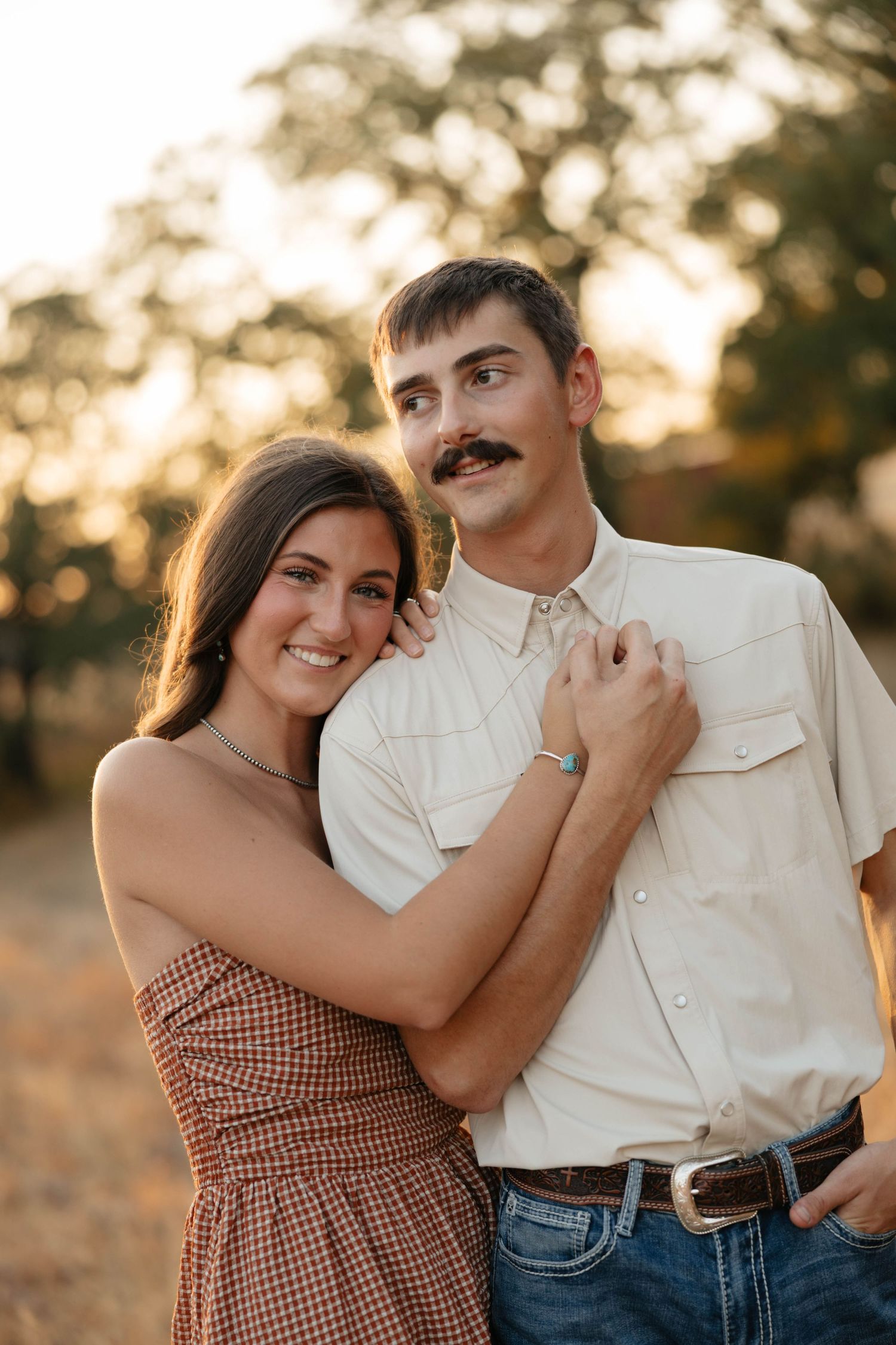 Smiling couple embracing at golden hour, man with mustache in western shirt and jeans.