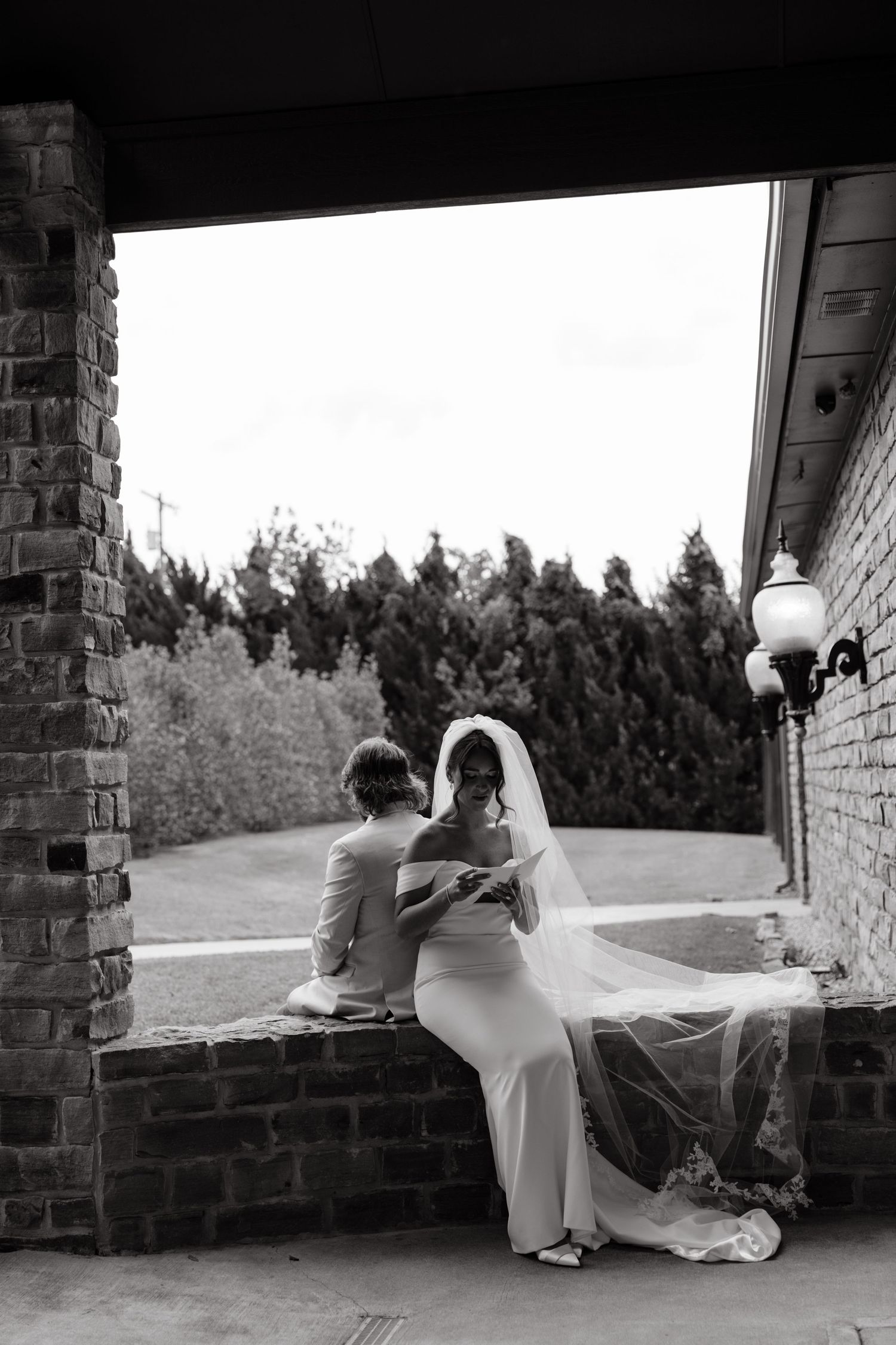 Black and white photo of bride in veil reading vows with groom on stone ledge at elegant outdoor wedding venue.