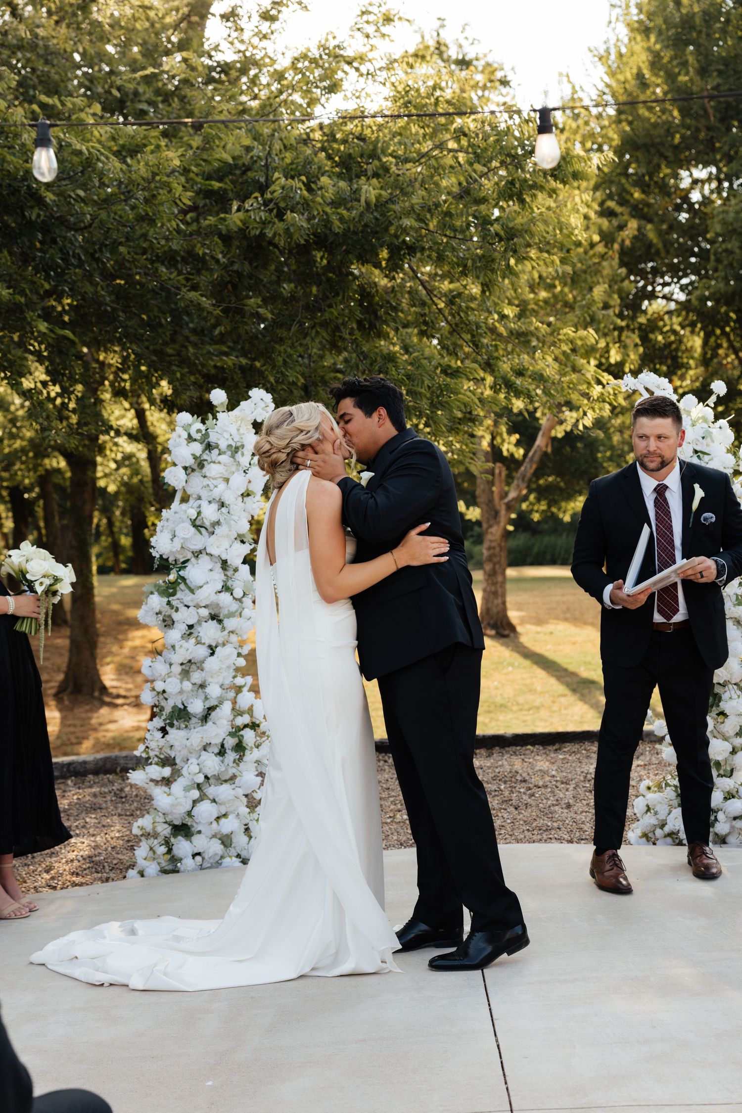 Bride and groom share first kiss at outdoor wedding ceremony before white floral arch at sunset.
