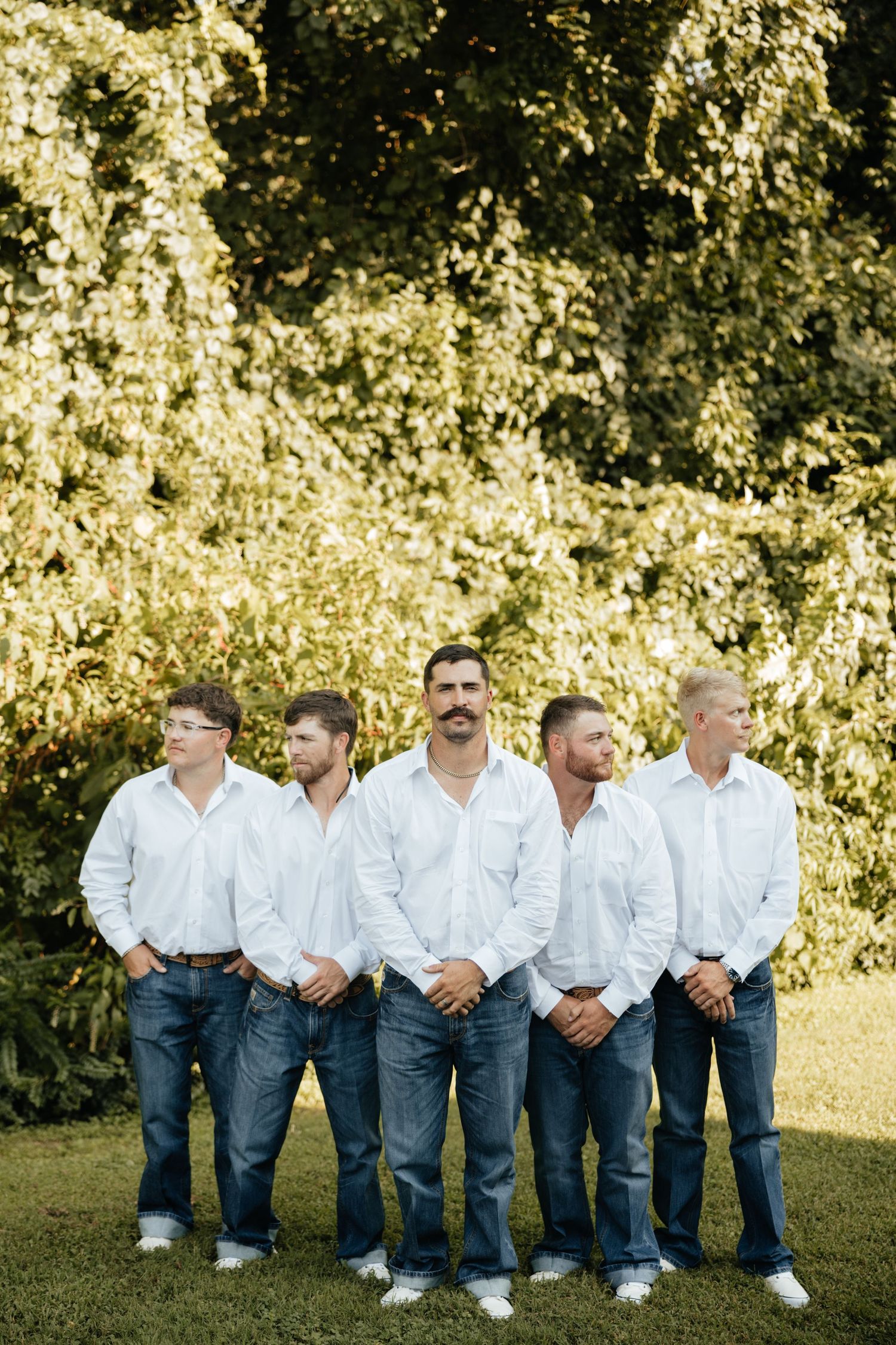 Five groomsmen in matching white shirts and jeans posing outdoors in front of lush green foliage.