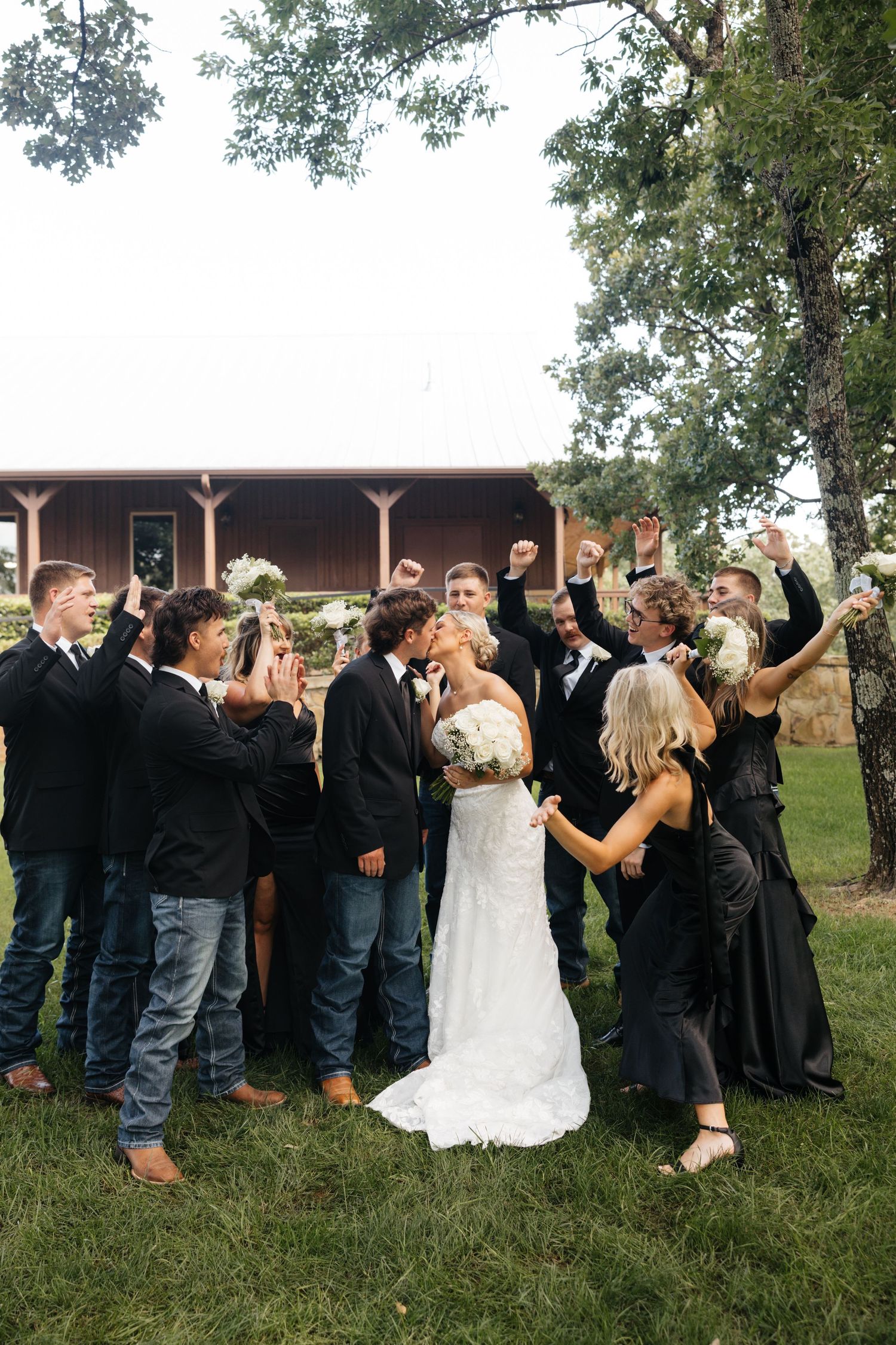 Wedding party celebrating newlyweds kissing outdoors near rustic barn venue surrounded by cheering guests.