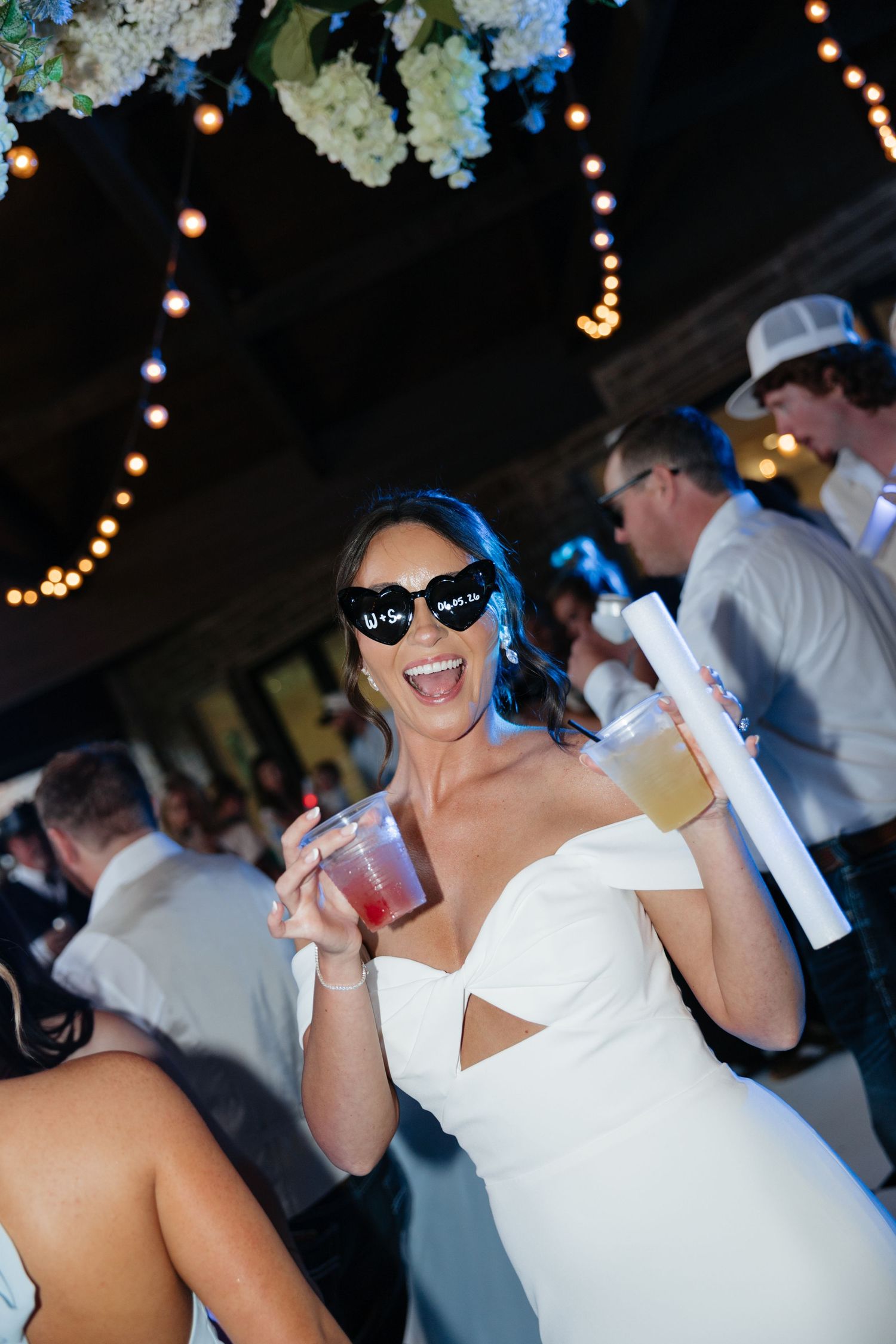 Joyful bride in white off-shoulder gown wearing Mrs. sunglasses holds two drinks while celebrating at her reception.