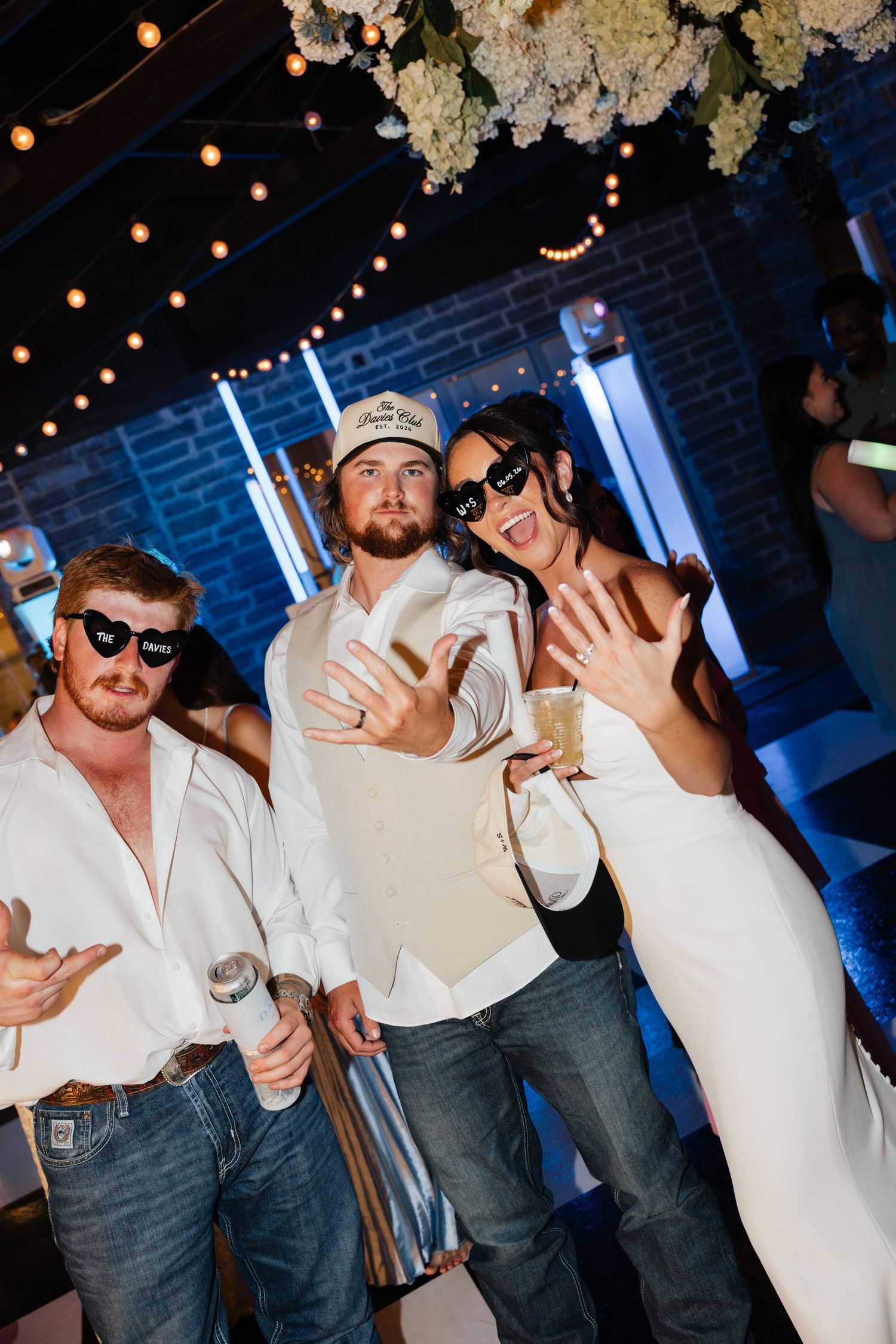 Three wedding guests pose playfully wearing sunglasses and fun accessories on the dance floor at a reception.
