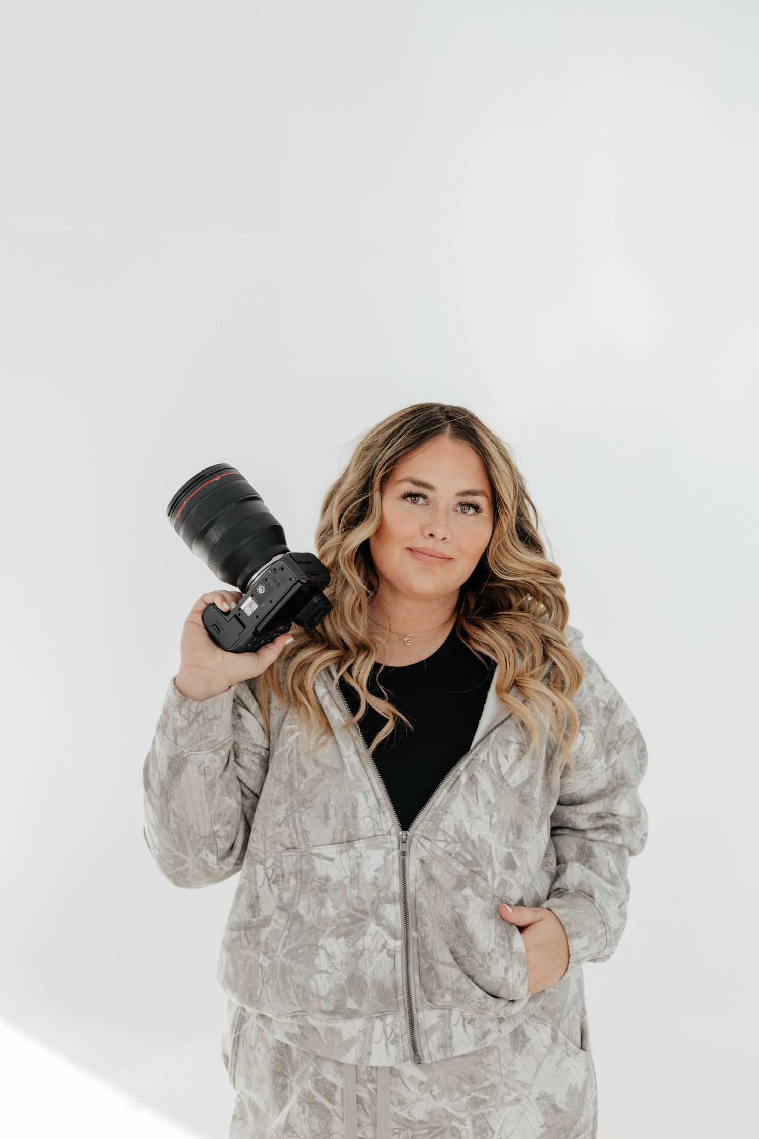 Female photographer holding a professional camera in a grey zip-up hoodie against white background.