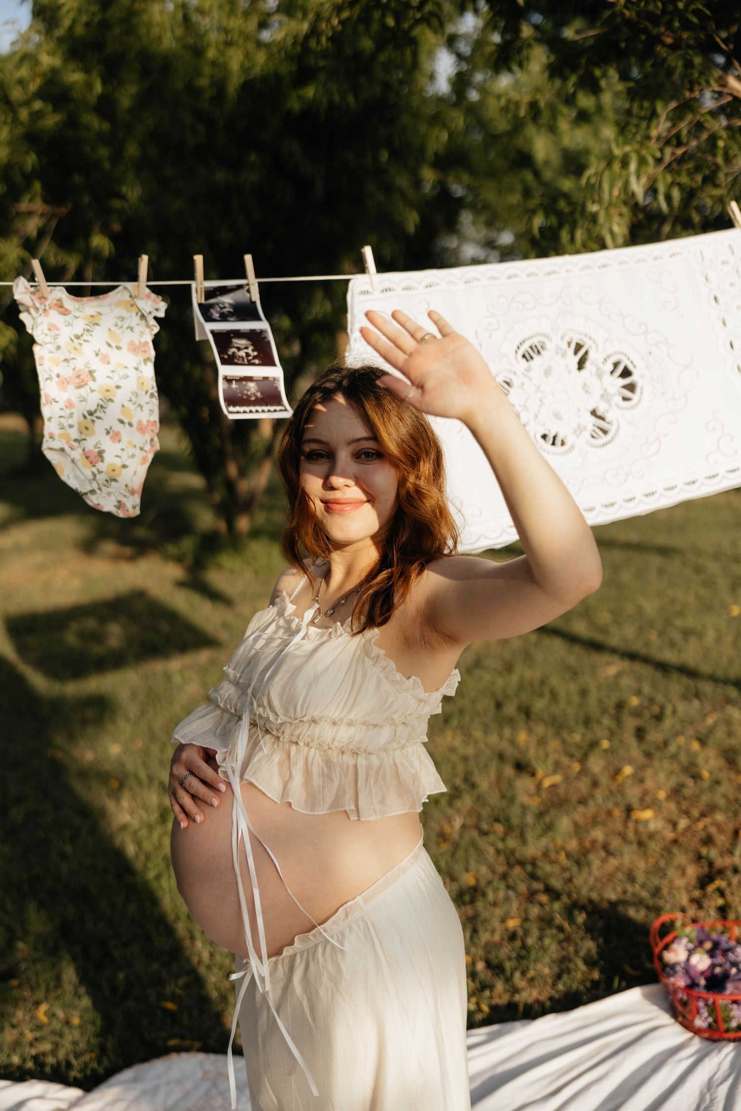 Pregnant woman in white crop top outdoors with ultrasound photos and baby clothes on a clothesline.