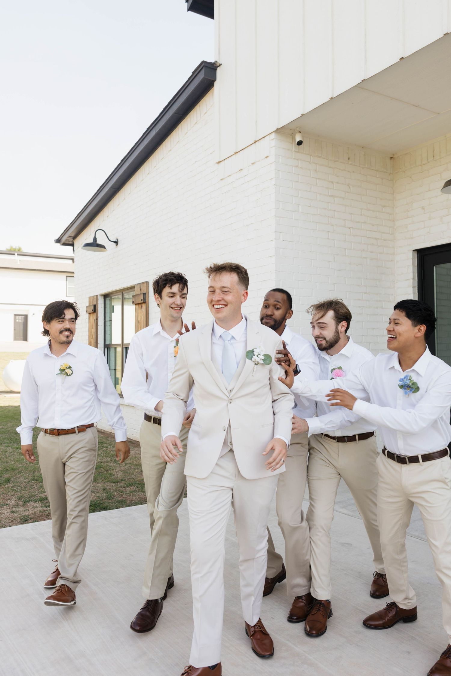 Groom in white suit walks with smiling groomsmen in khaki pants and white shirts outside a white brick wedding venue.