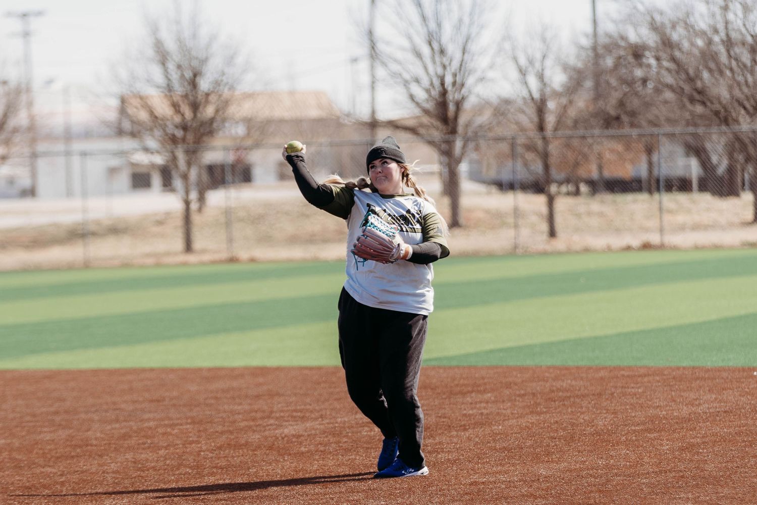 Female softball player throwing ball on an outdoor dirt field on a sunny day.