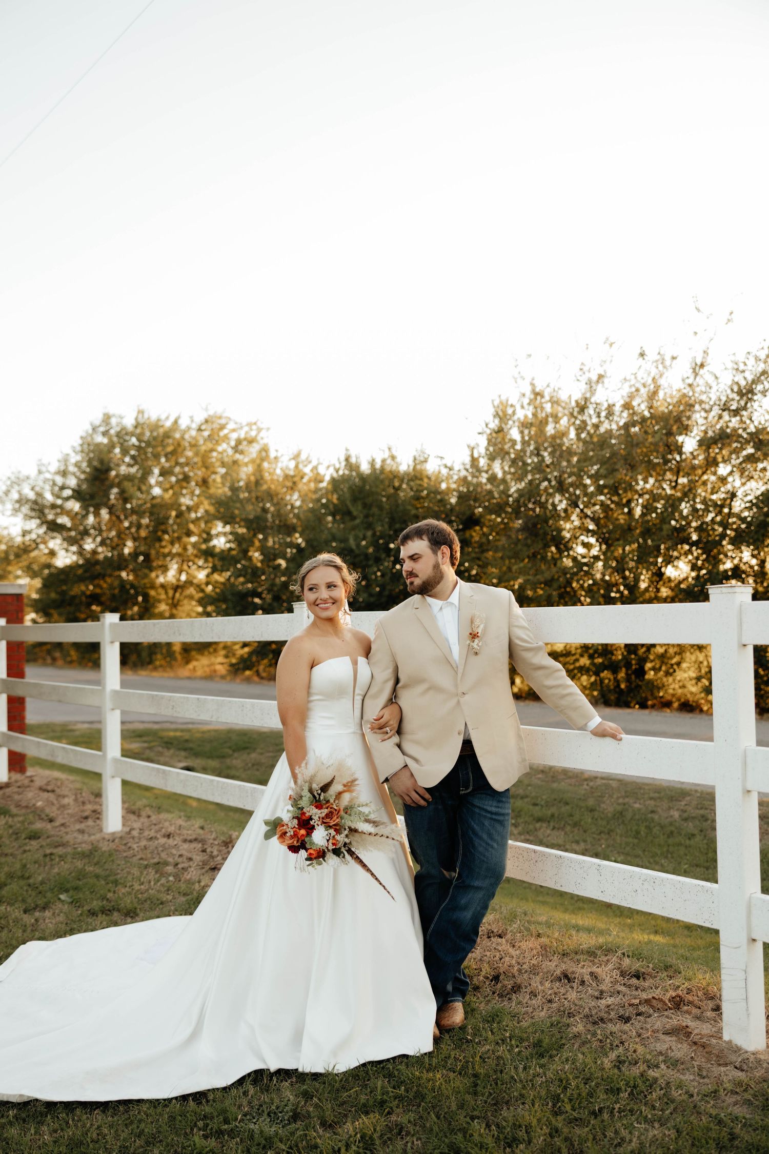 Bride and groom posing by a white fence at golden hour, bride holding a colorful bouquet.