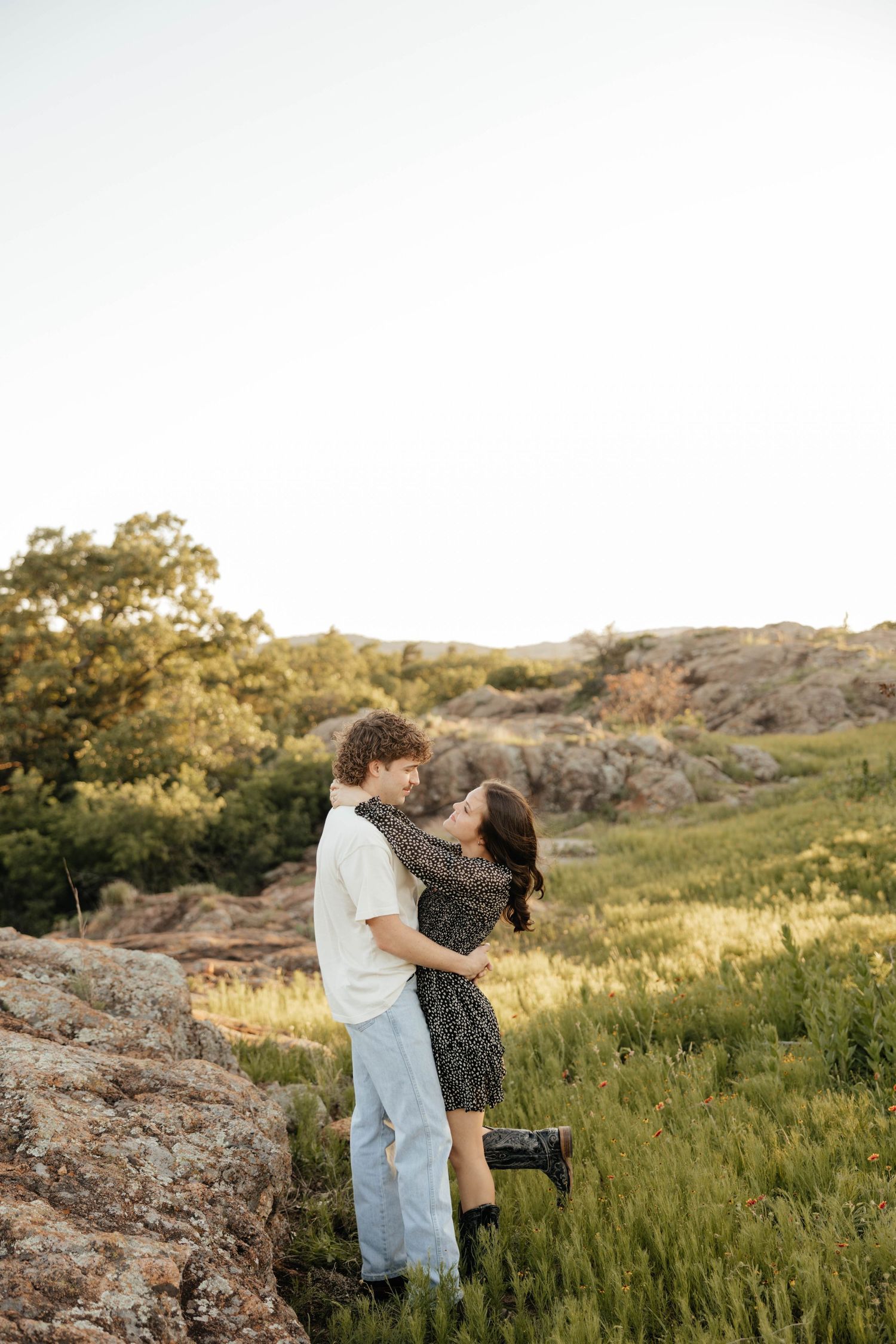 Couple embracing and laughing together on rocky hillside during golden hour engagement session outdoors.