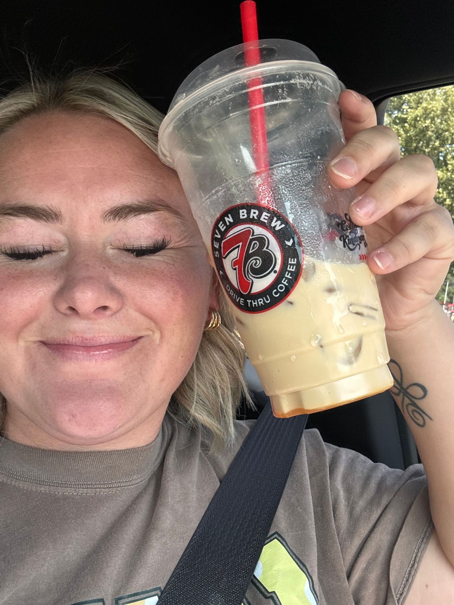 Smiling blonde woman holding an iced coffee from 7 Brew Drive-Thru Coffee in a car.