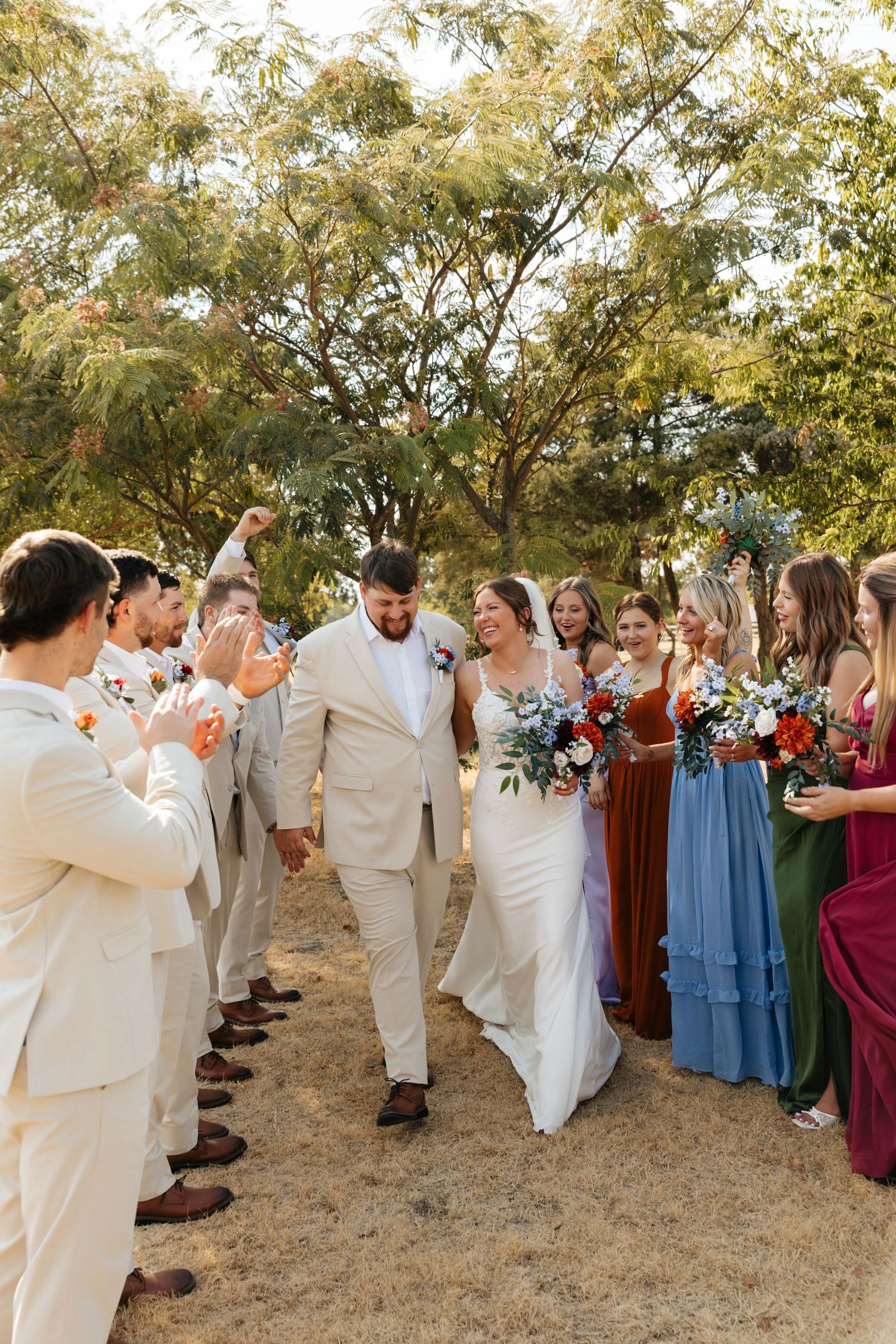 Bride and groom walk through cheering wedding party in colorful mismatched bridesmaid dresses and tan suits outdoors.