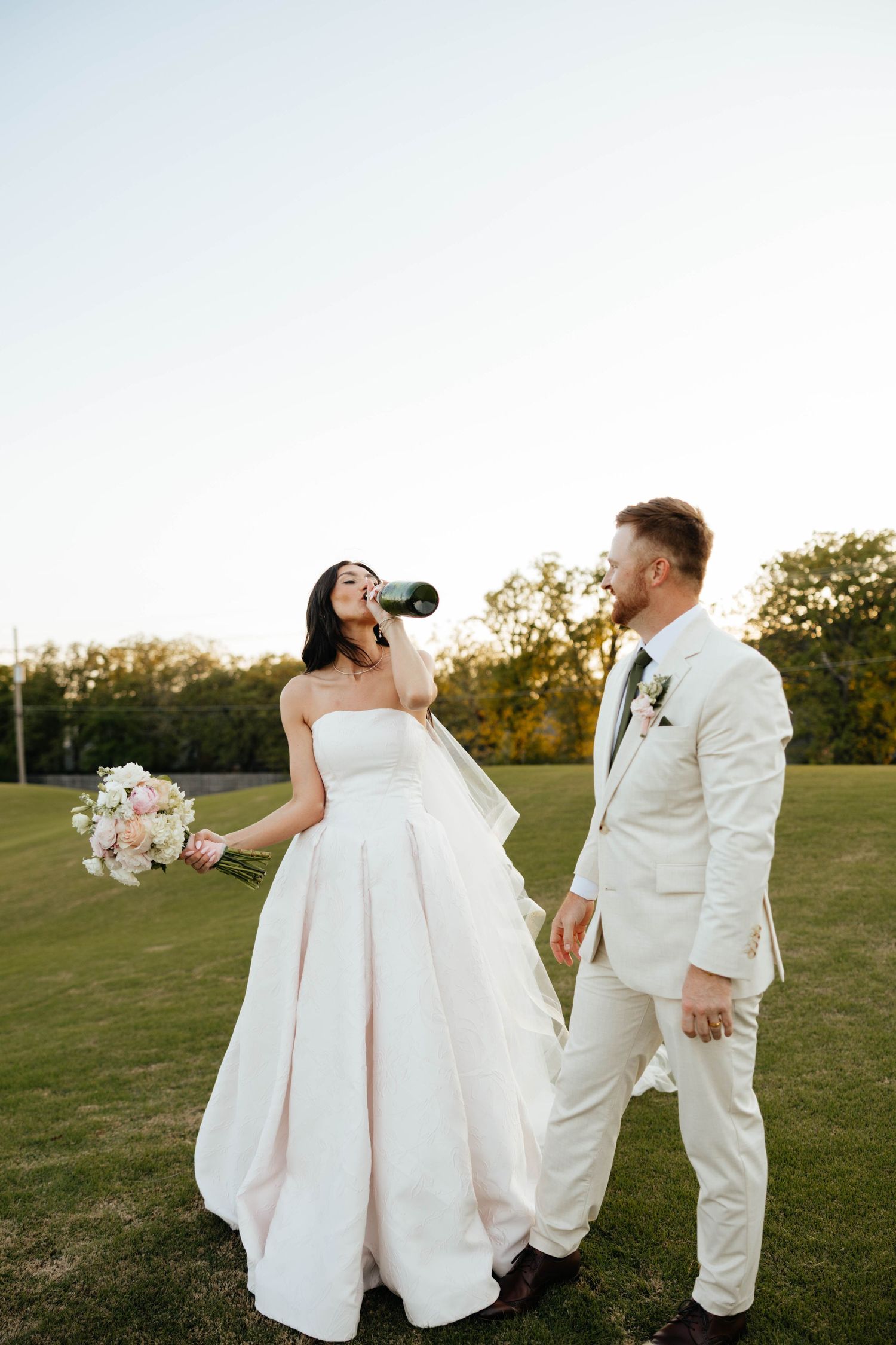 Bride drinks champagne from bottle in blush gown while groom in ivory suit holds her hand on green lawn.