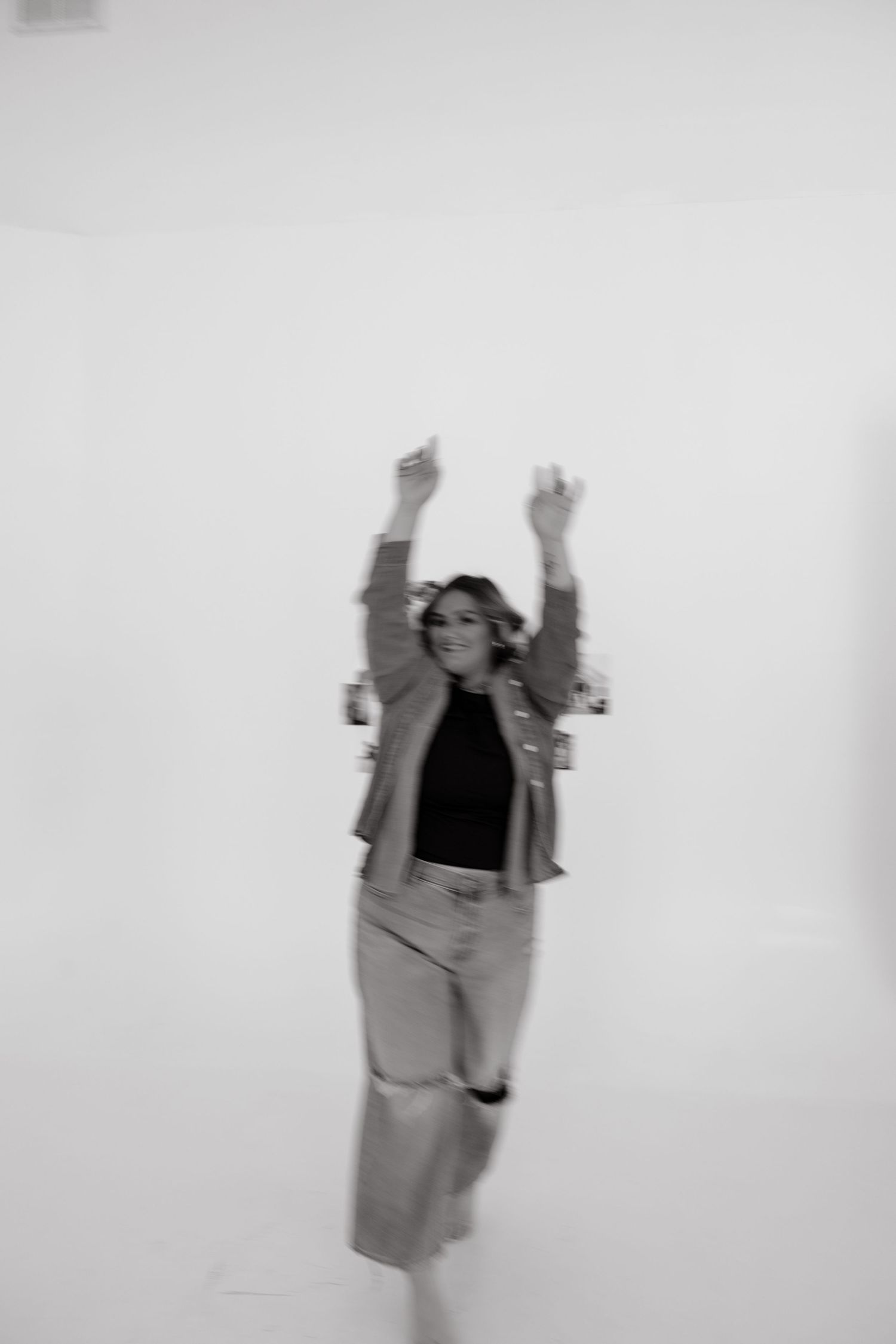 Black and white photo of a young woman in jeans and jacket dancing with arms raised in a bright white studio.