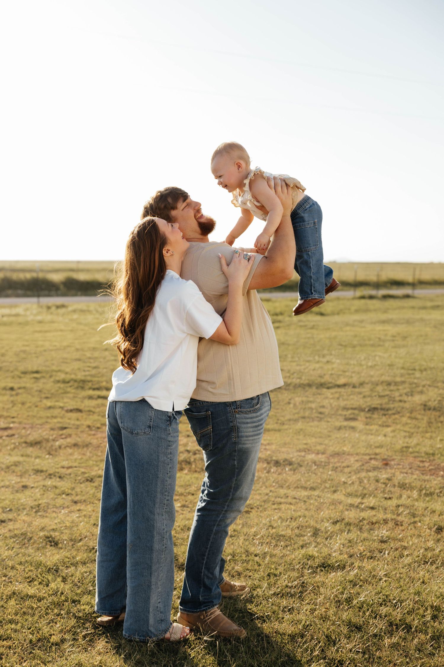 Happy young family with father lifting laughing baby in sunny green field at sunset.