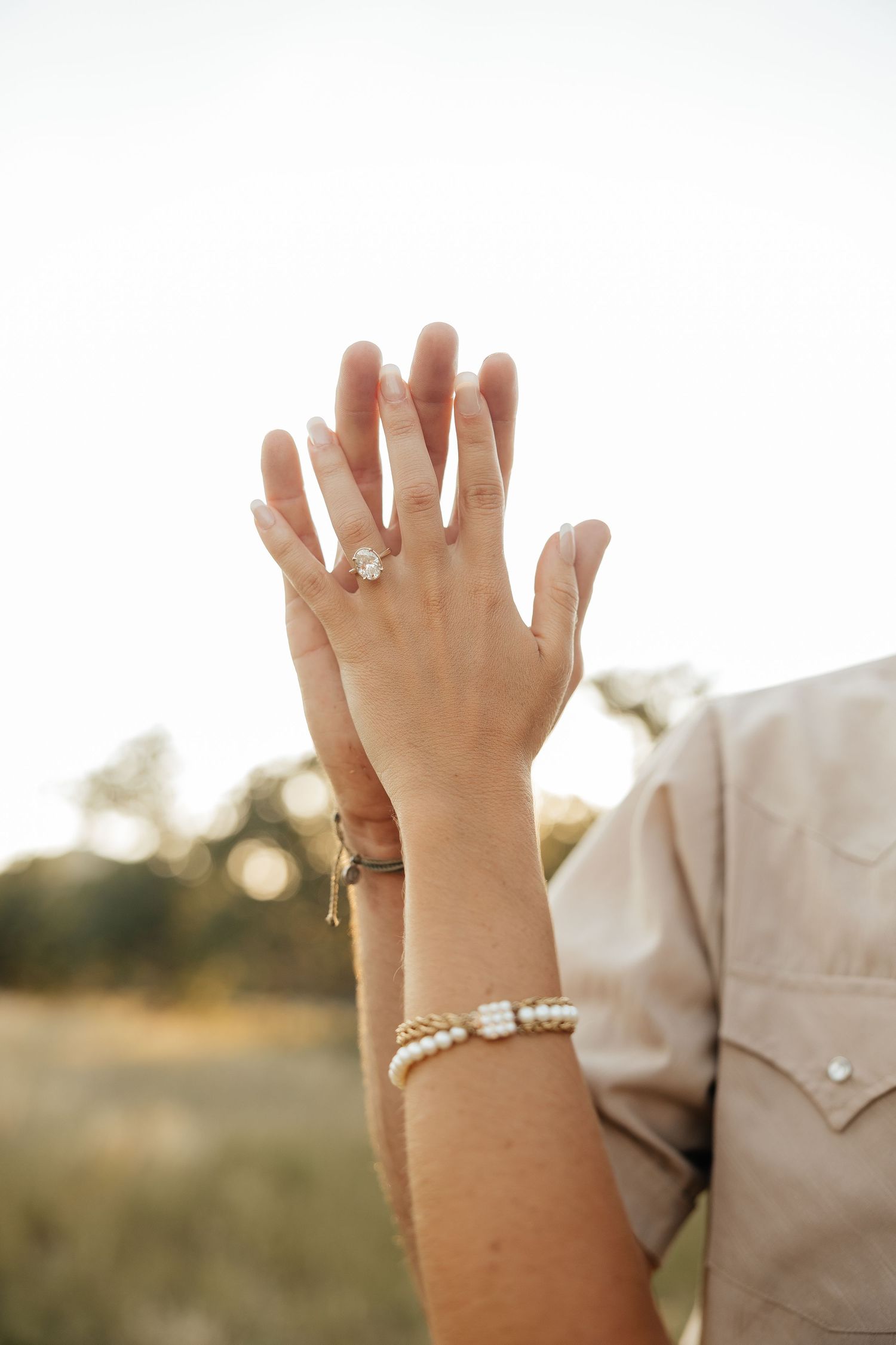 Close-up of engaged couple holding hands up showing diamond engagement ring and pearl bracelet outdoors.