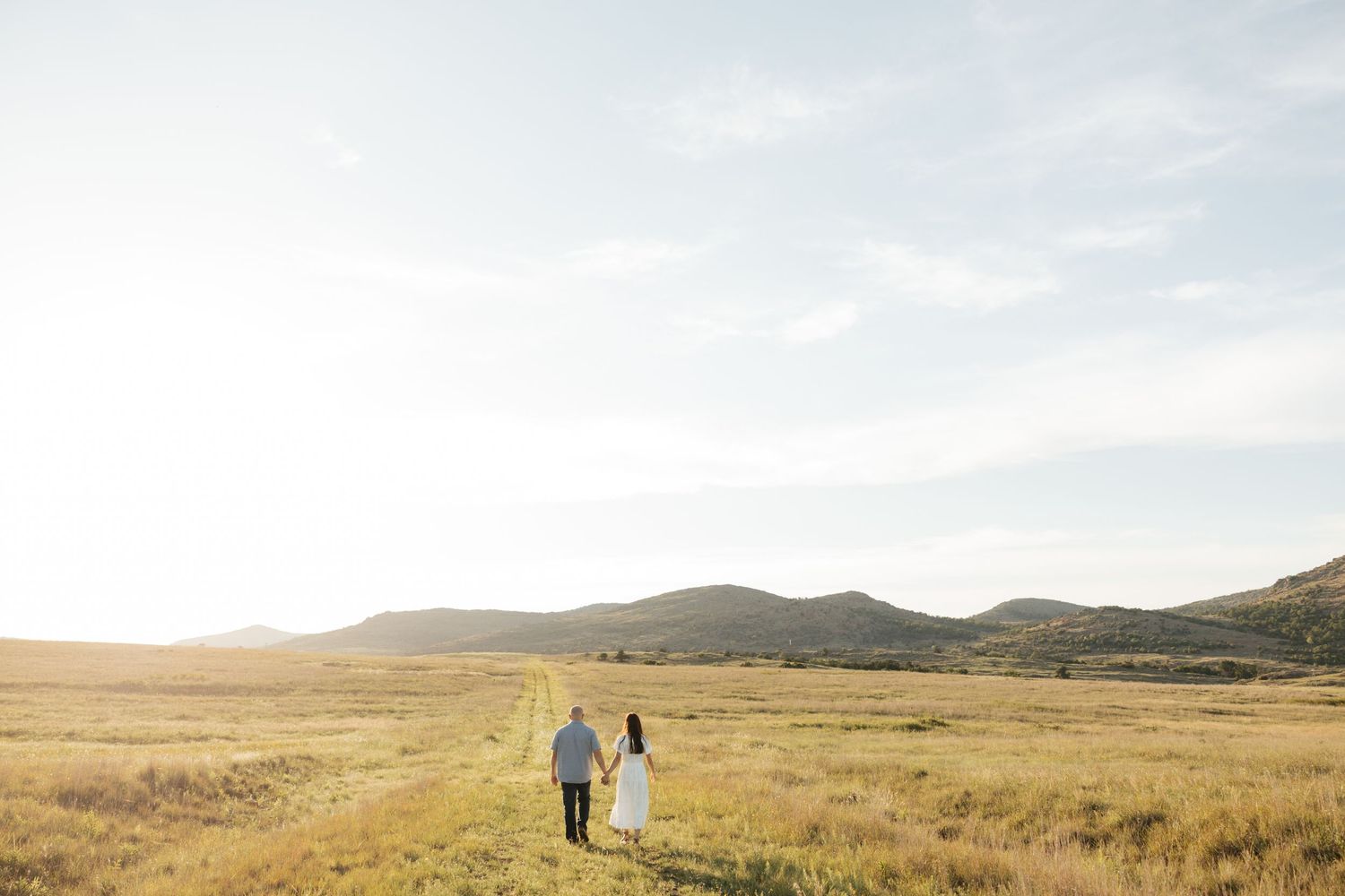 Couple holding hands walking through golden meadow with mountain backdrop at sunset.