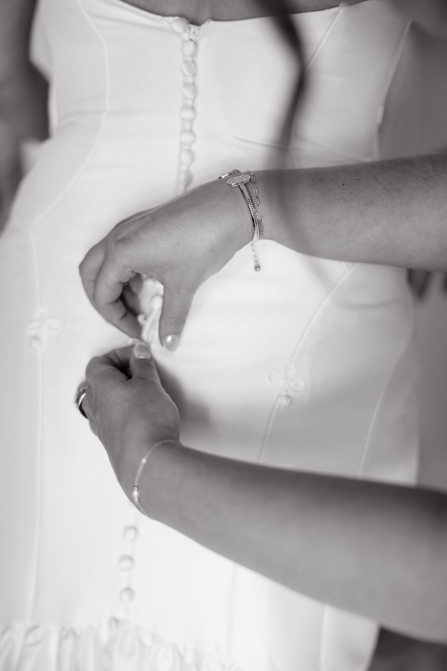 Black and white close-up of hands buttoning the back of a wedding dress with delicate buttons.