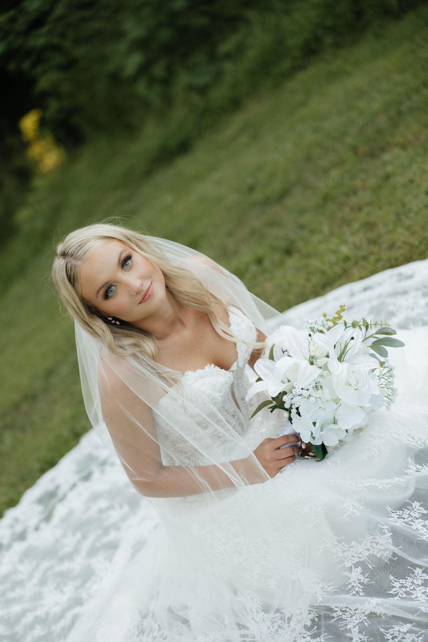 Blonde bride in lace gown and veil sits on grass holding white floral bouquet, gazing upward outdoors.