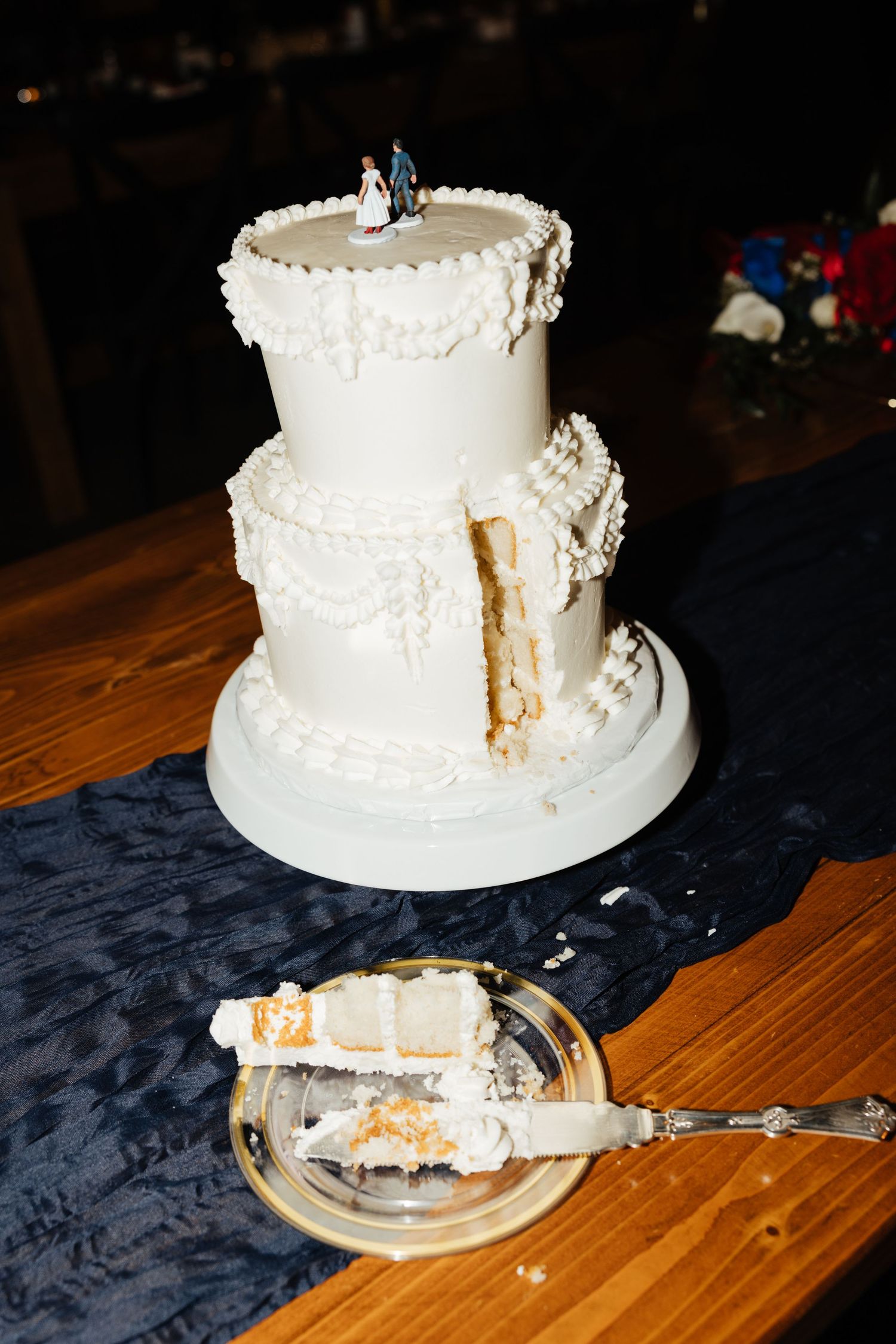 Two-tier white pearl-decorated wedding cake with bride and groom topper on navy table runner.