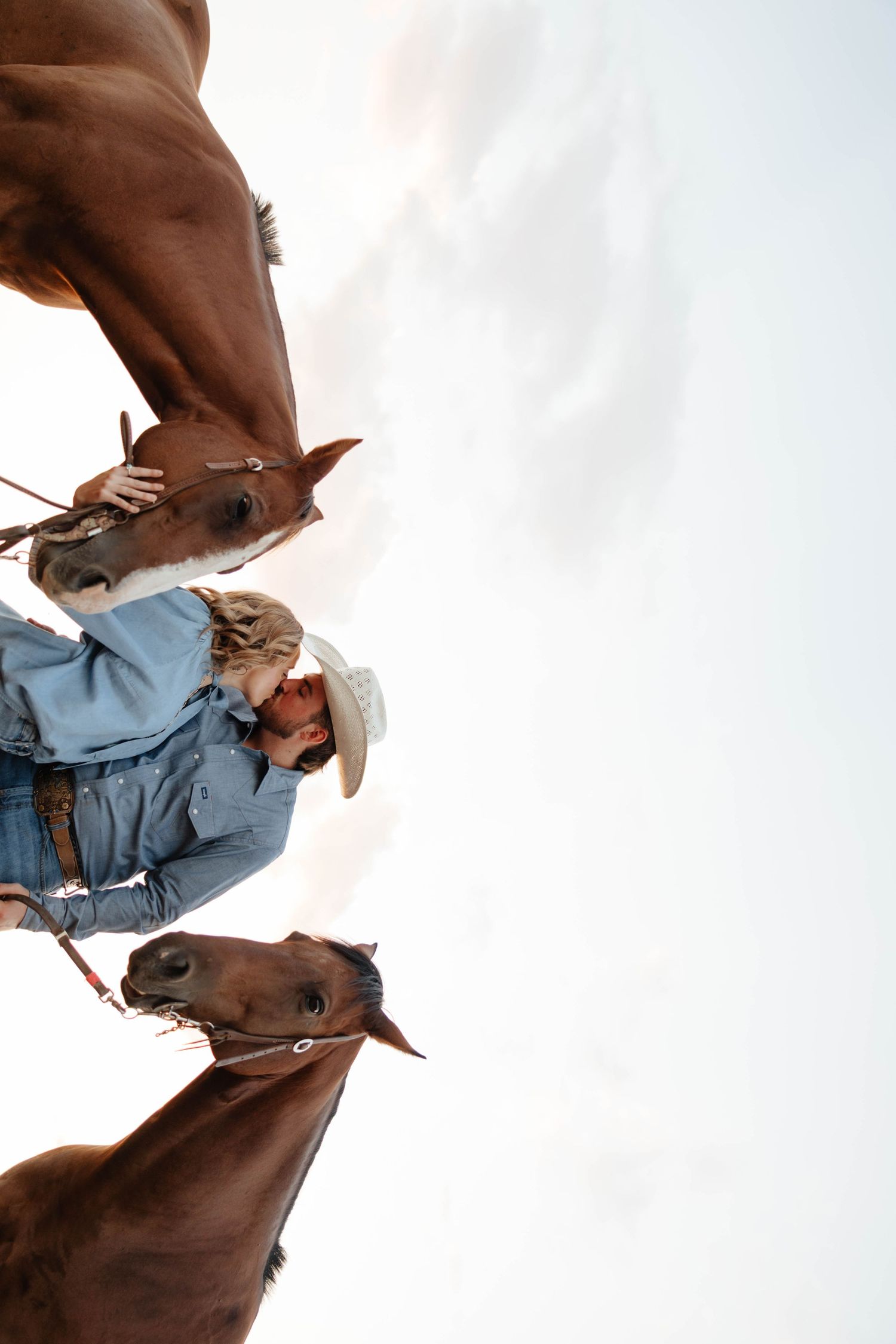 A cowgirl in a white hat stands between two chestnut horses against a bright sky.