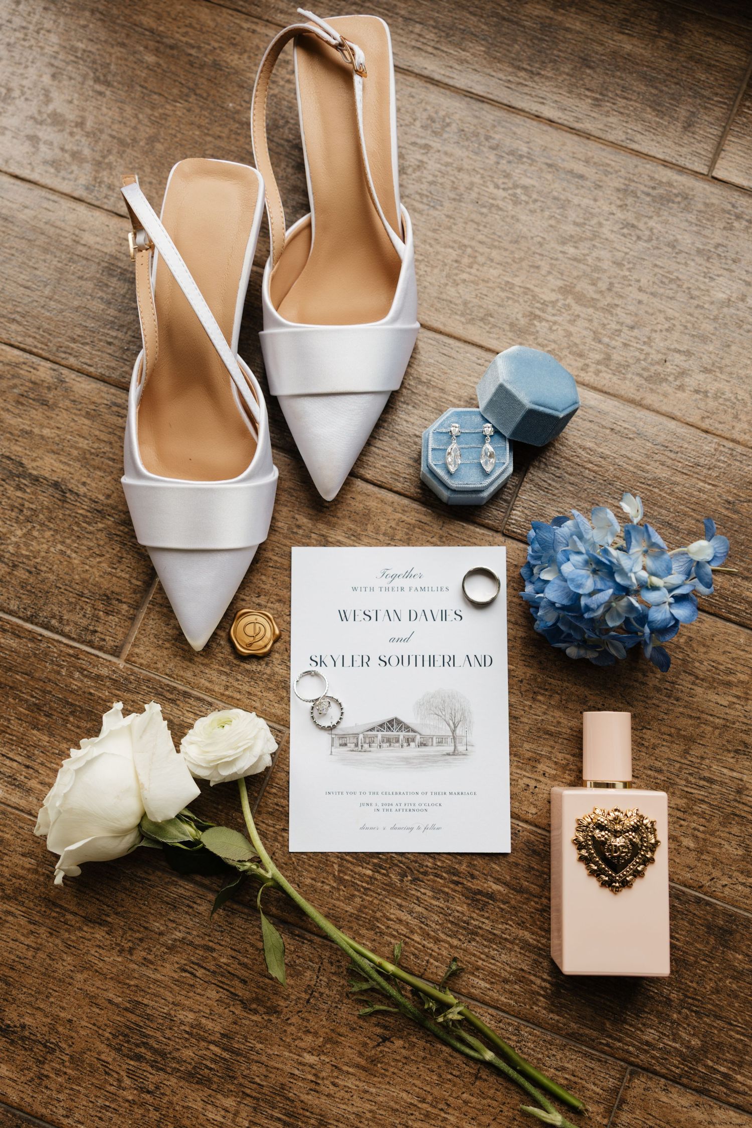 A flat lay of white wedding heels, invitation, rings, earrings, perfume, and blue flowers.