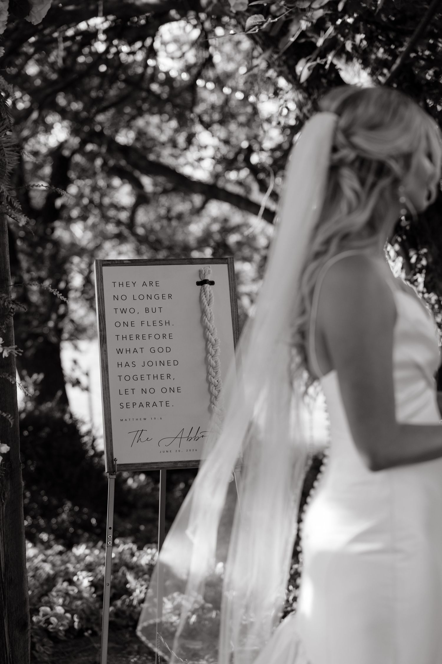 Black and white photo of bride standing near Matthew 19:6 scripture sign at wedding ceremony.