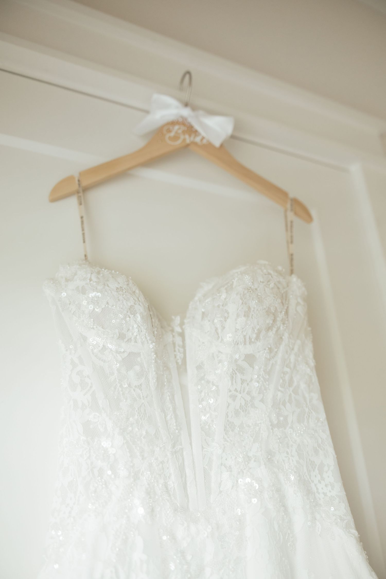 Elegant beaded strapless wedding dress hanging on wooden bridal hanger against white wall.