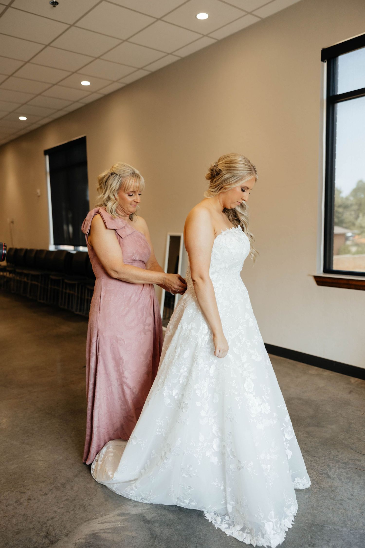 A bride in a white lace gown being helped by a woman in a dusty rose dress in a bright modern room.