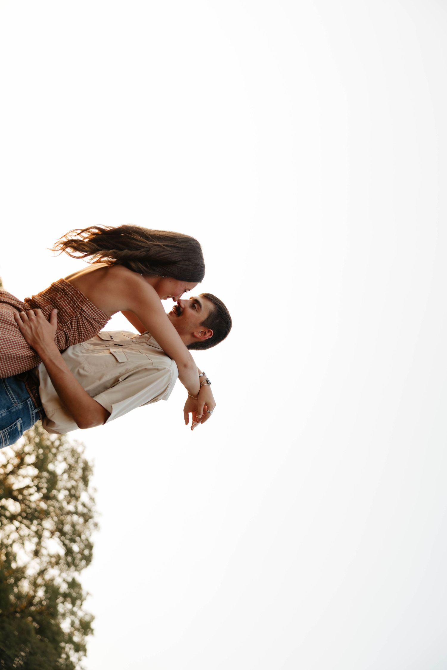 Man lifting woman for a kiss outdoors, captured from below against a bright sky.