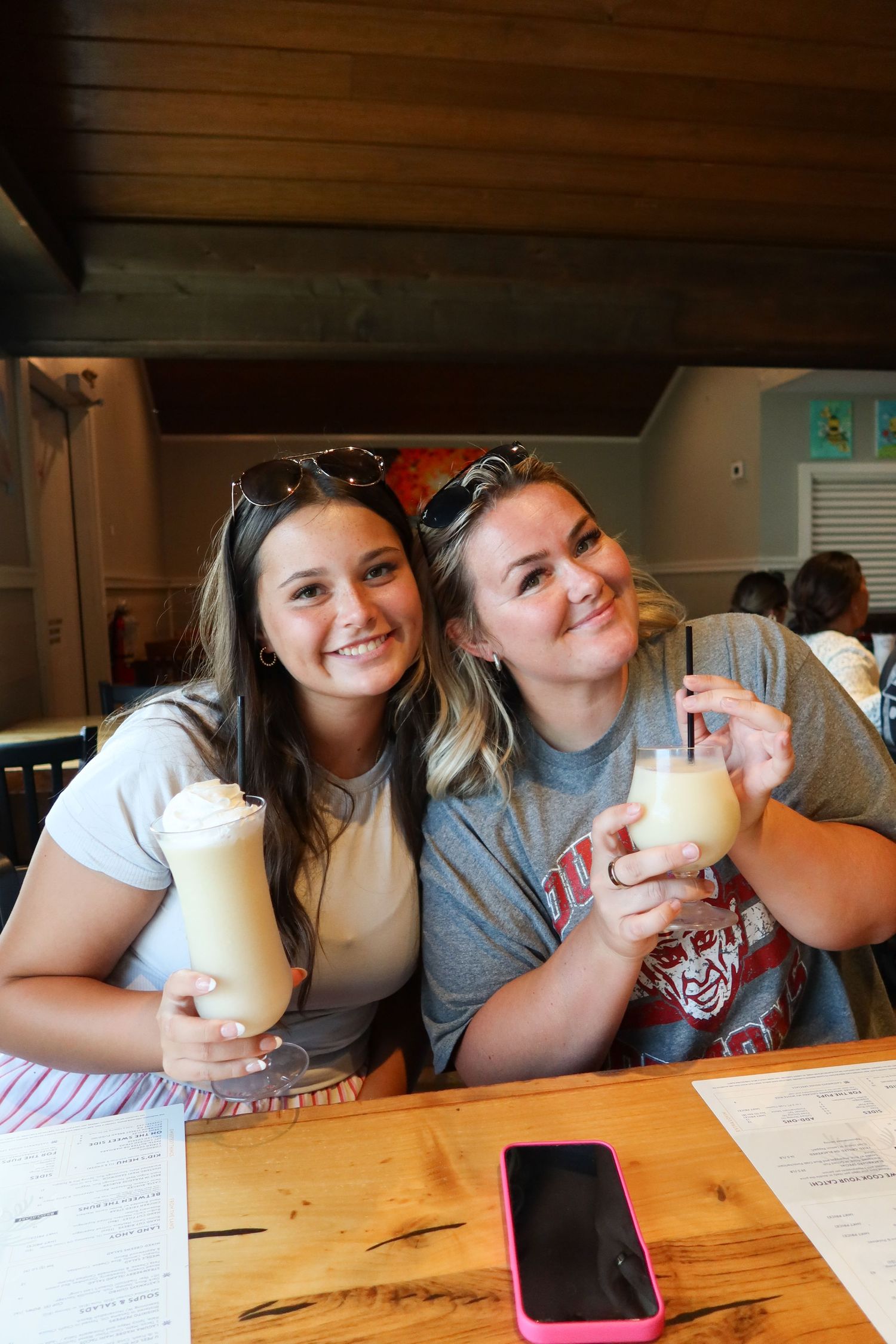 Two young women smiling and holding creamy cocktails at a restaurant table.