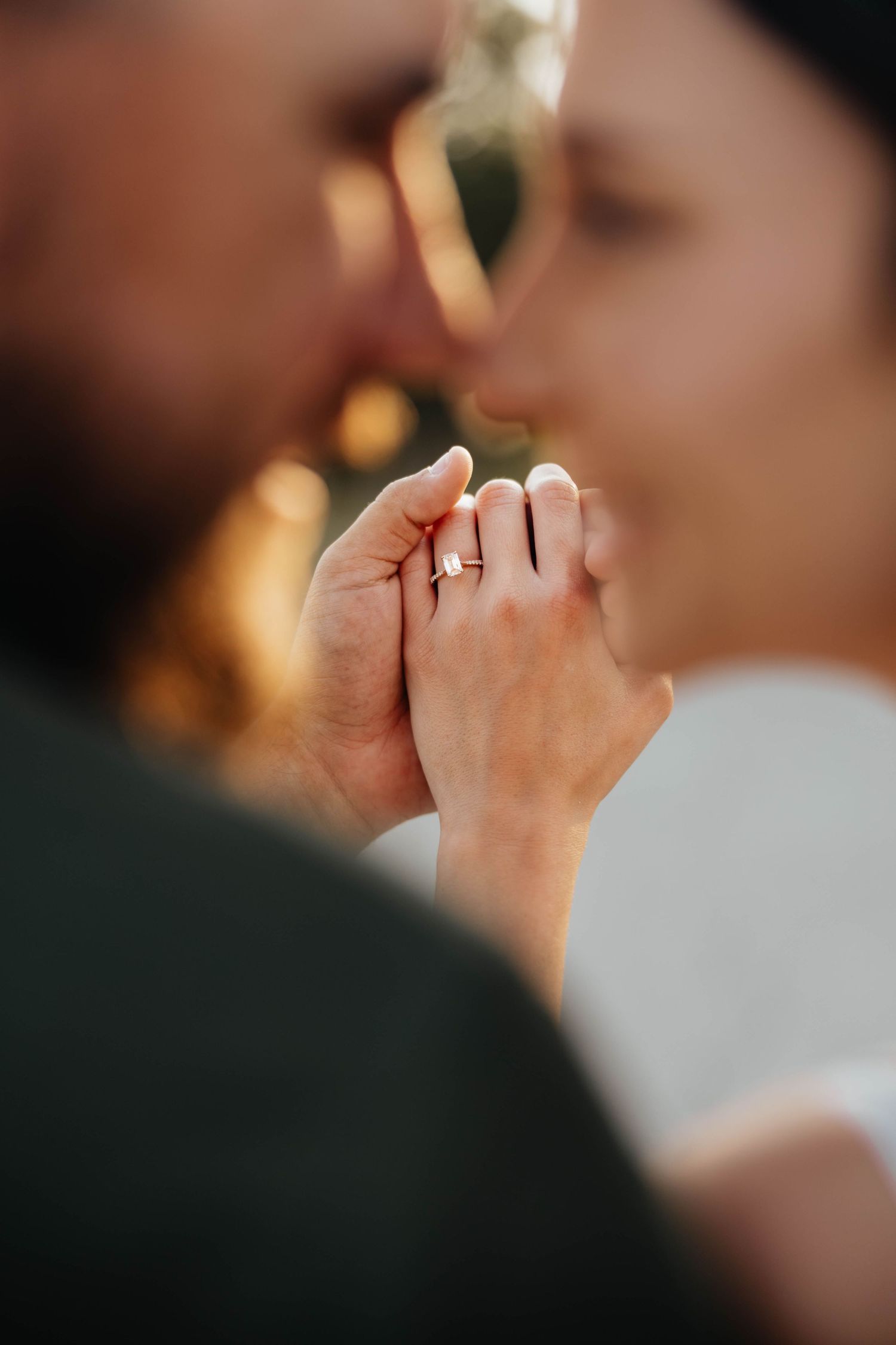 Close-up of couple kissing while holding hands showing an engagement ring in soft bokeh light.