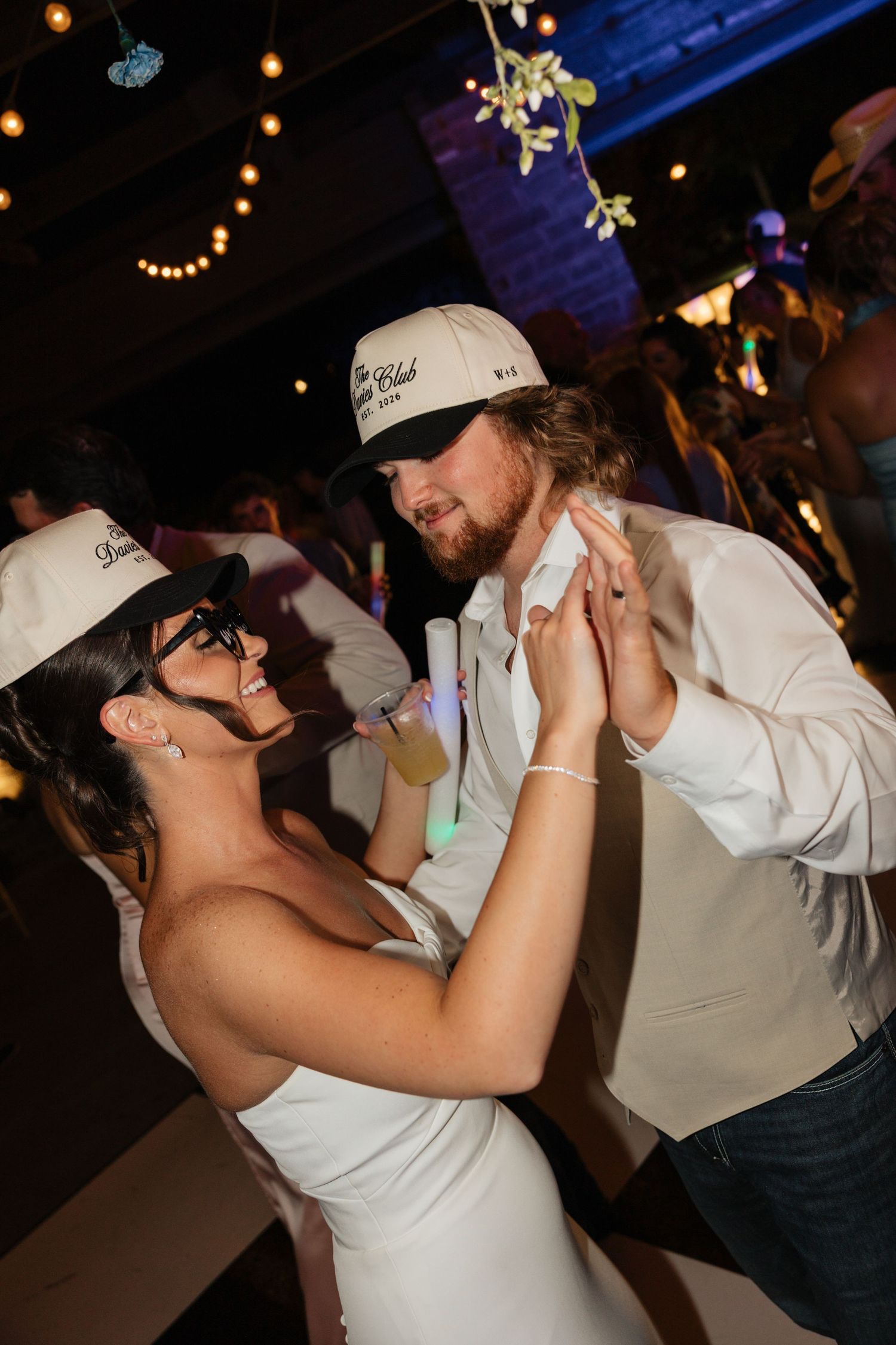 Bride and groom dance together wearing matching Davies Club trucker hats and glasses at their wedding reception.