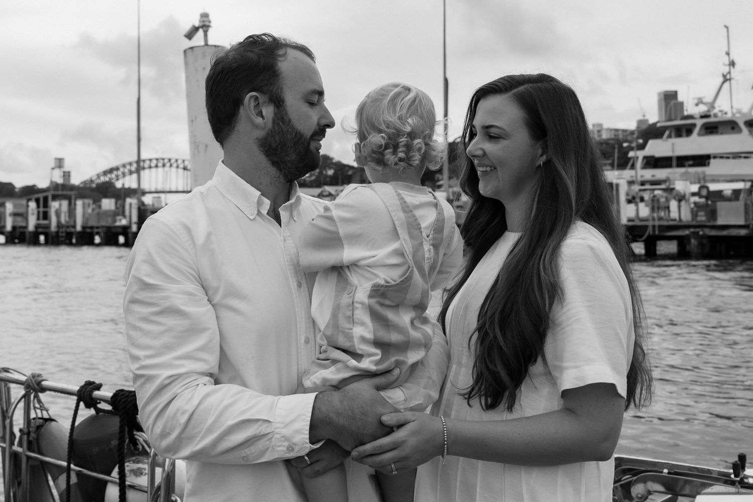 Black and white photo of a couple holding a toddler on a harbour waterfront with a bridge in the background.