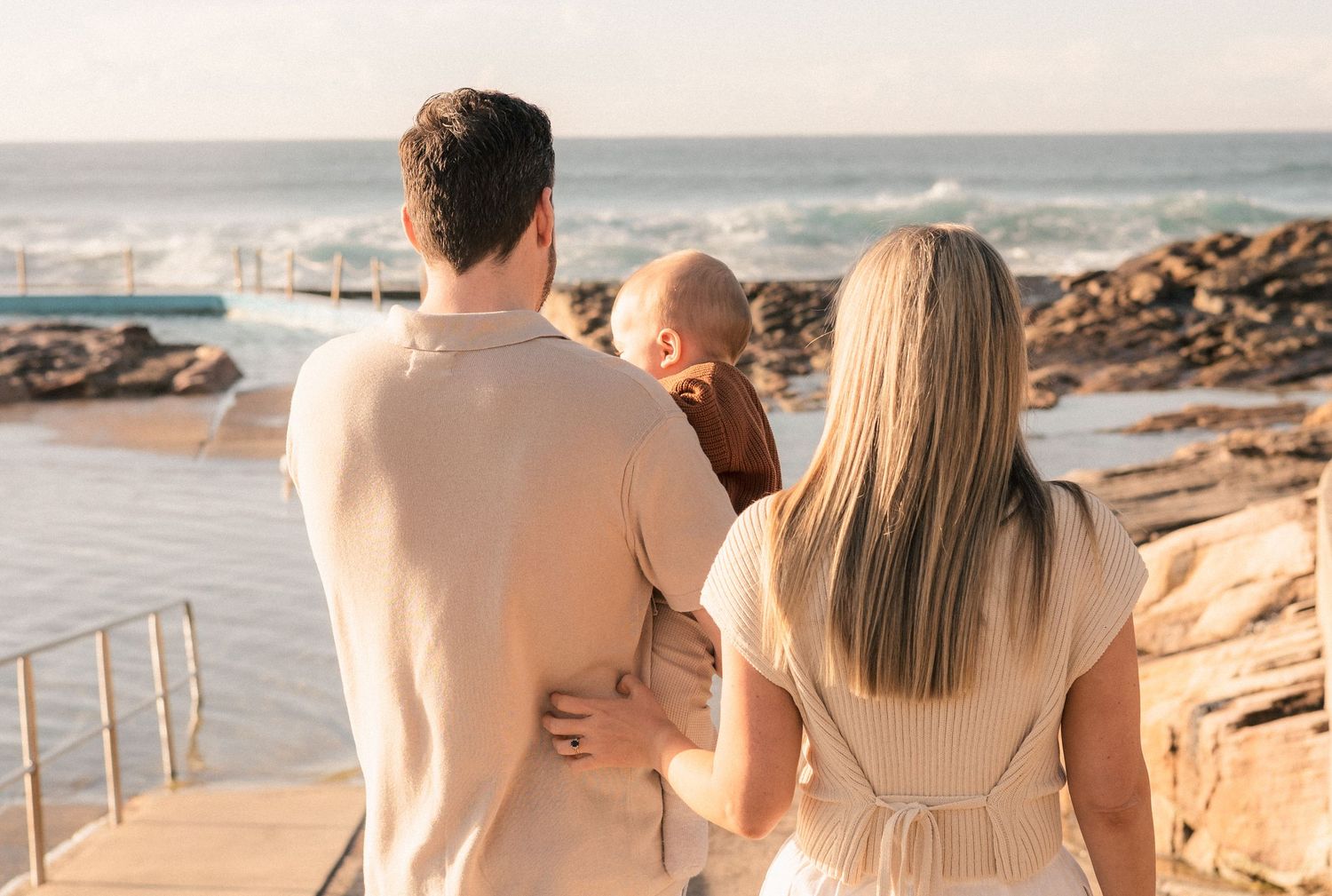 A family of three stands together facing the ocean, with parents holding their baby near a coastal rock pool.
