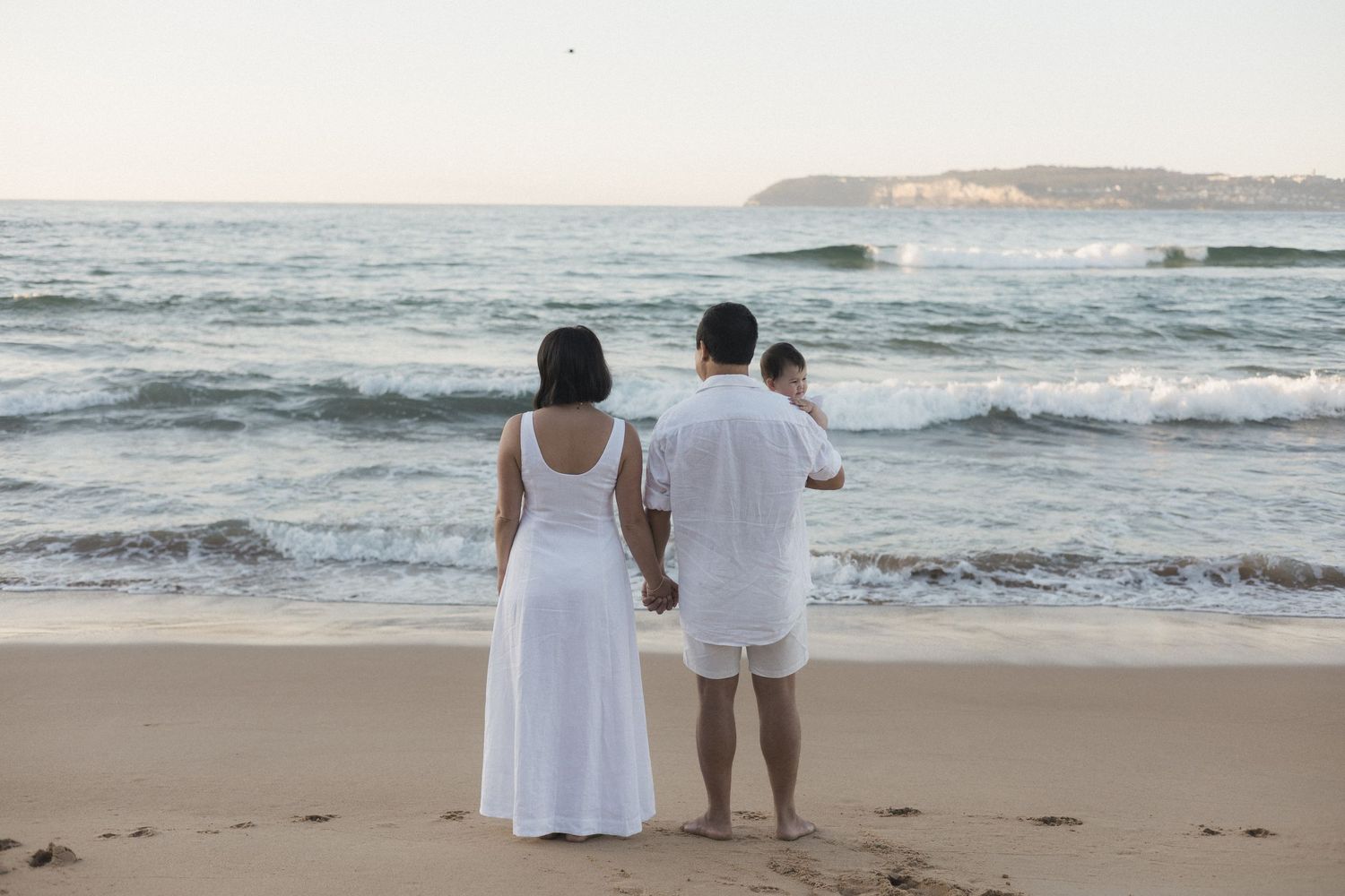 Family in white outfits standing on sandy beach holding hands, gazing at ocean waves at dusk.