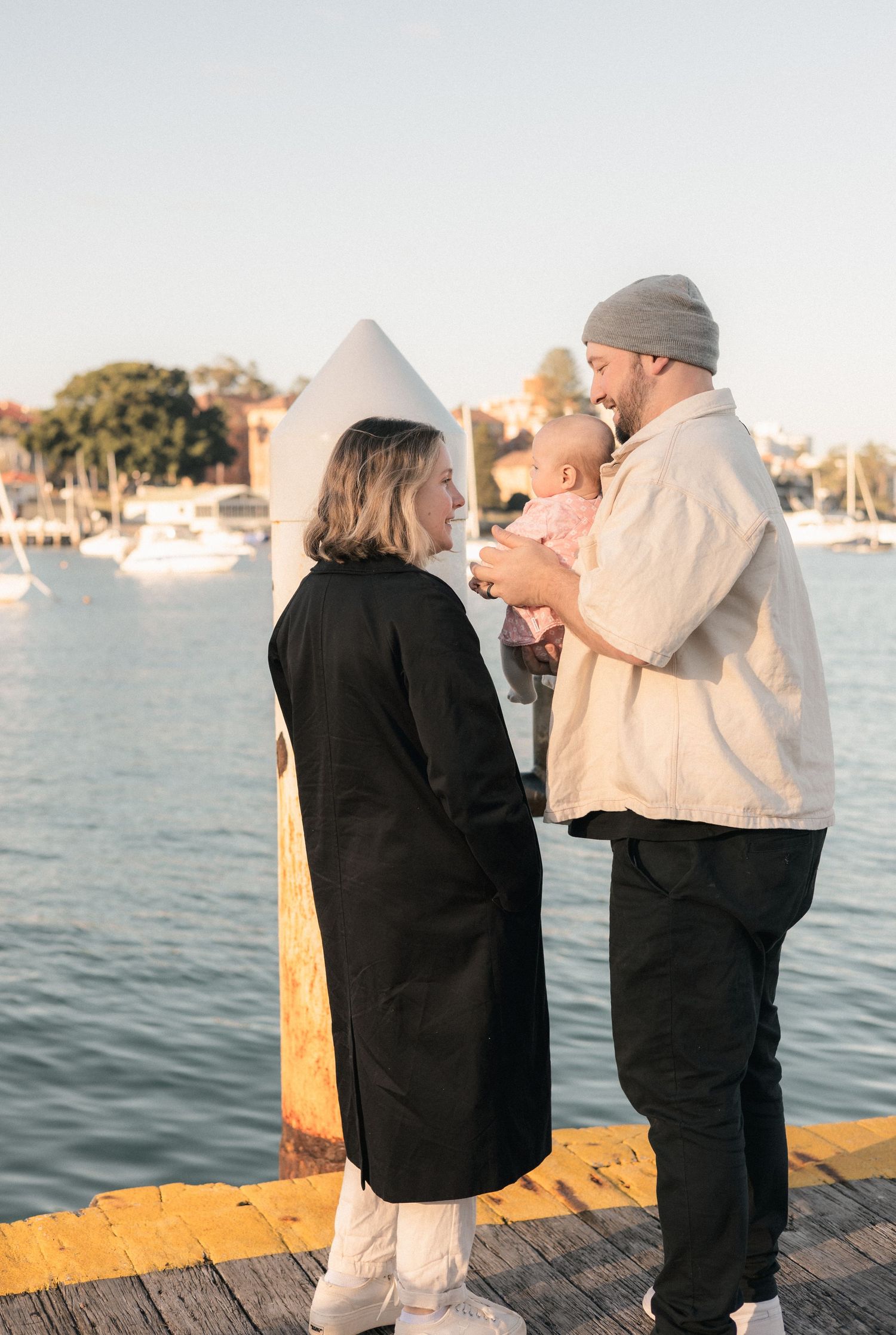 A couple stands on a sunny dock holding their baby, with sailboats and waterfront homes visible behind them.