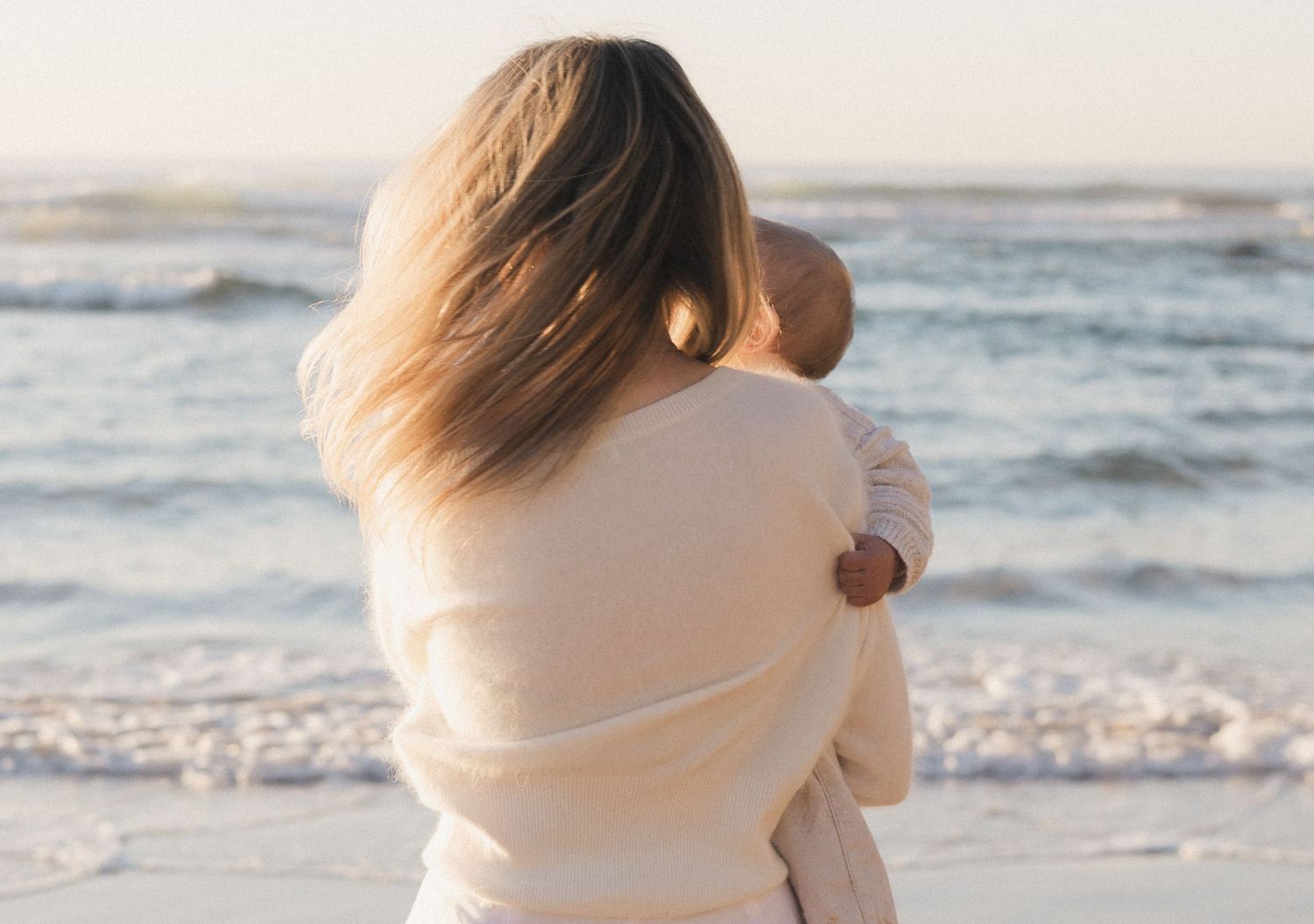 A mother holding her baby on a serene beach, both dressed in cream, facing the calm ocean waves.